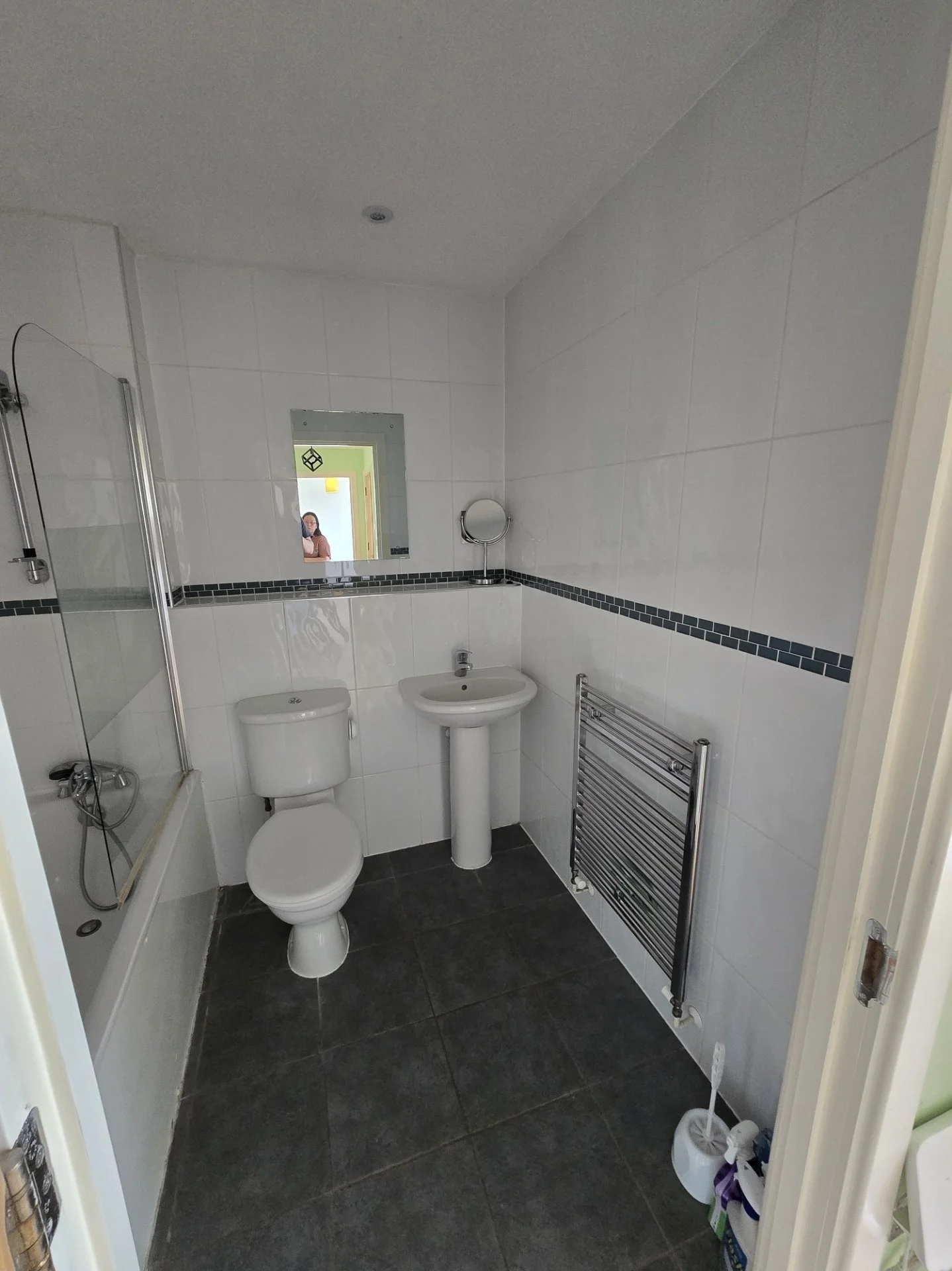 A small bathroom with white tiled walls and black tiled floor. It has a toilet, a pedestal sink, a mirror, and a shower with a glass door. There is a heated towel rail on the wall, and some bathroom cleaning items on the floor.