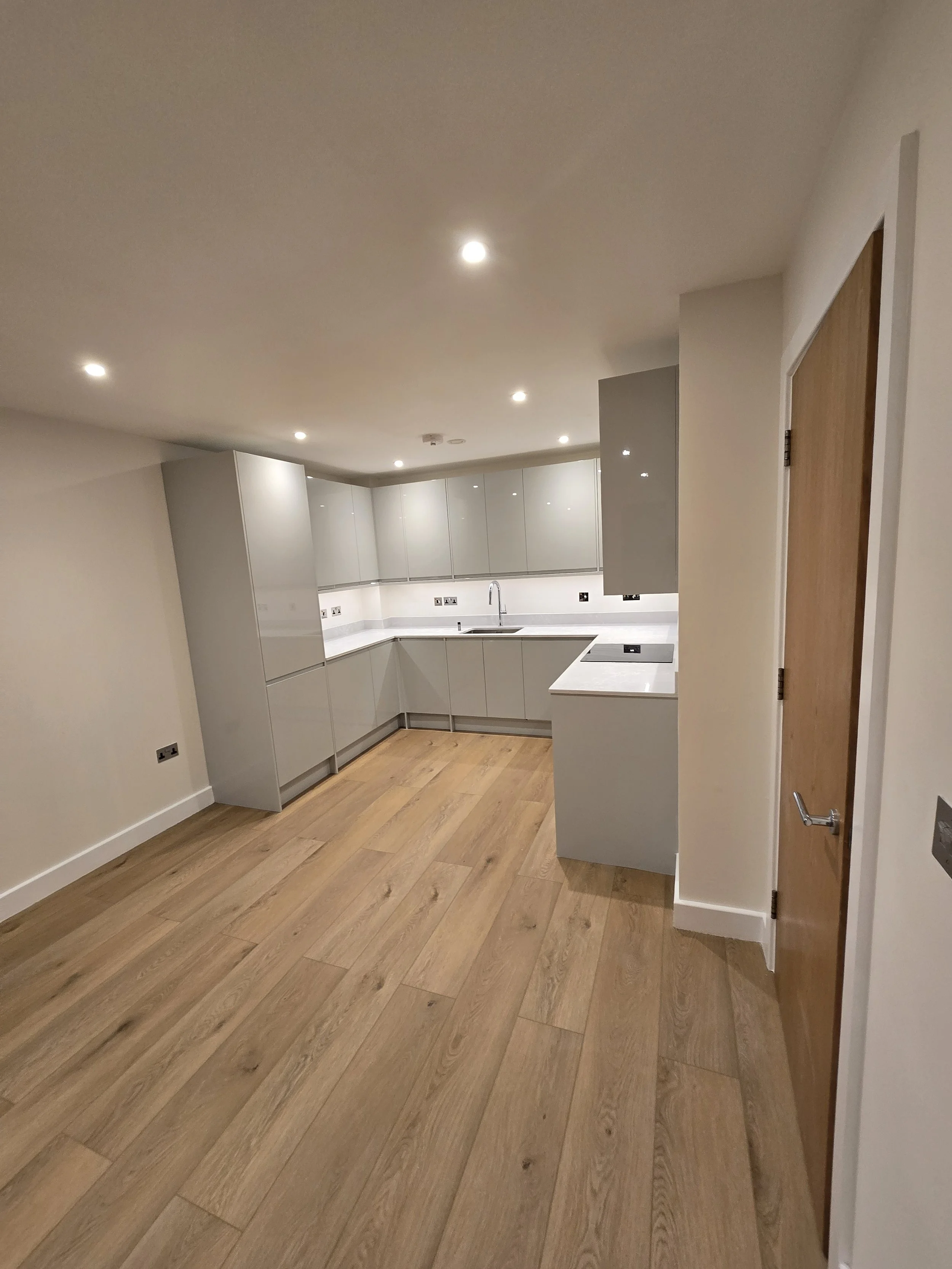 Modern kitchen with wooden flooring, white cabinets, a marble countertop, and a stainless steel sink with a gooseneck faucet. Recessed ceiling lights and a wooden door are visible.