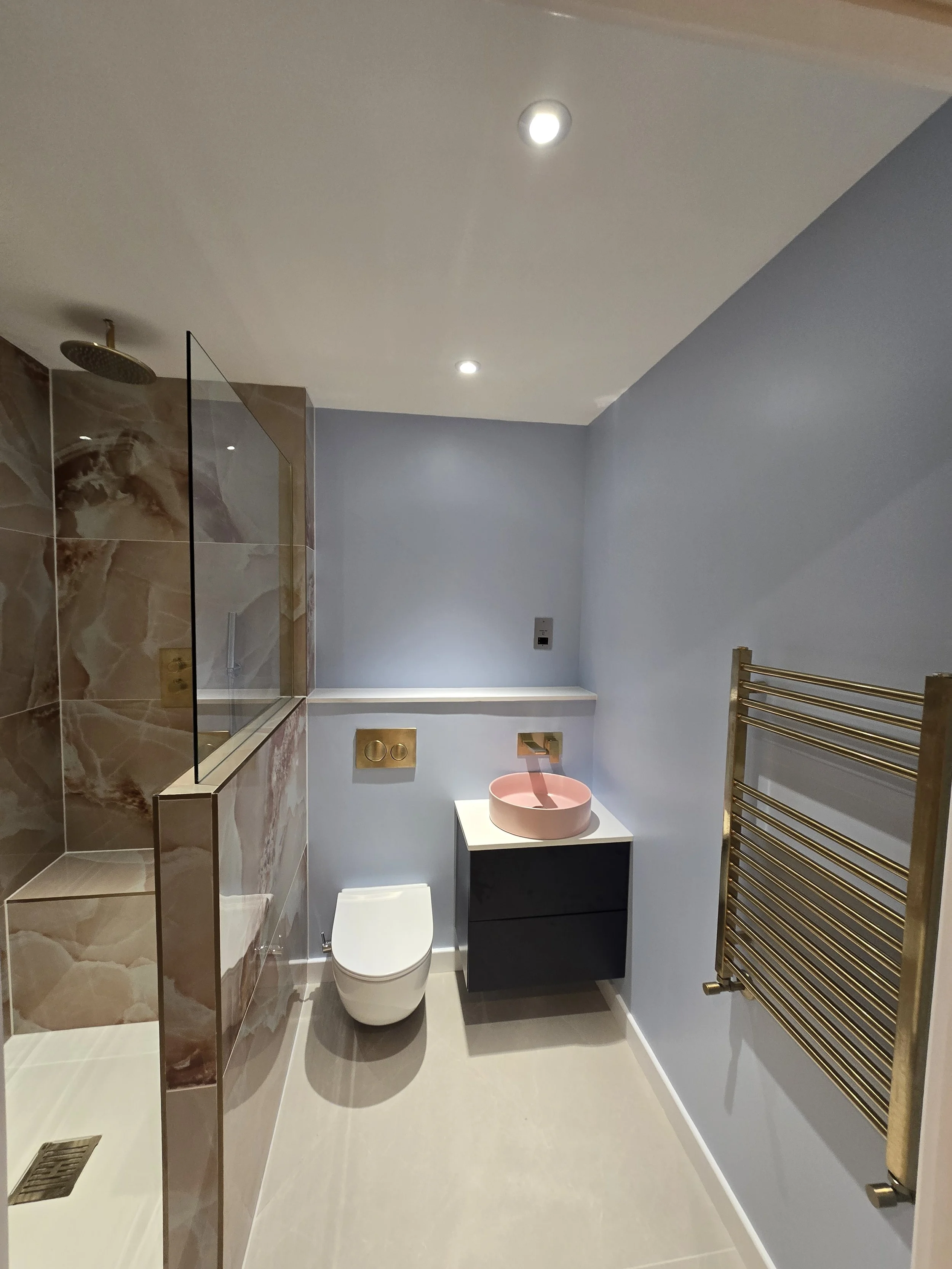 Modern bathroom with a walk-in shower with a marble wall, a wall-mounted toilet, a black vanity with a pink vessel sink, a wall-mounted toilet paper holder, a gold towel warmer, and a light blue painted wall with a small mirror.