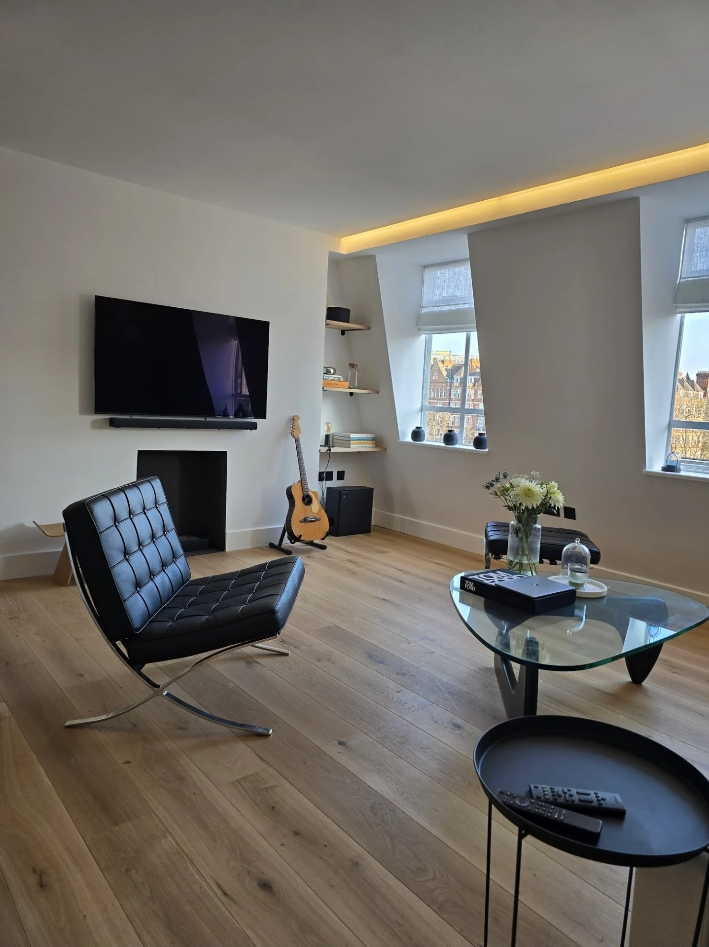 Modern living room with black leather chair, glass coffee table with books and a vase of flowers, flat-screen TV on white wall, guitar against wall, windows showing buildings outside, wooden flooring.