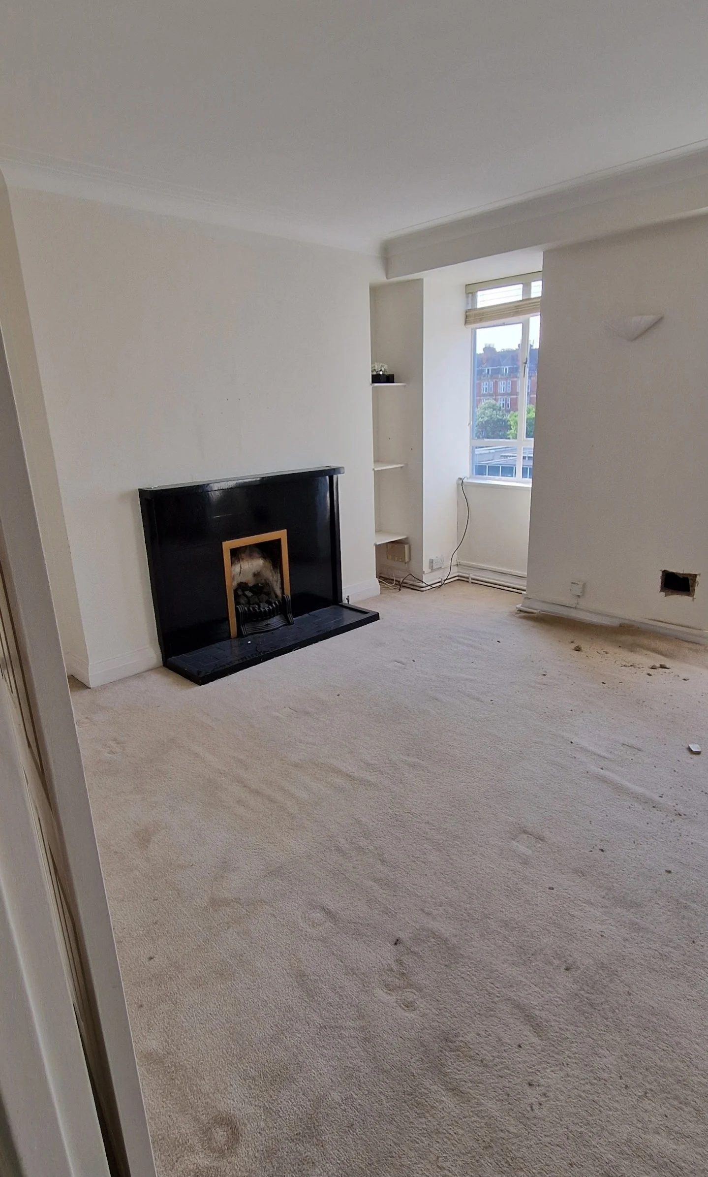 Empty living room with fireplace, large window with blinds, built-in shelves, and beige carpet with some dirt and debris.