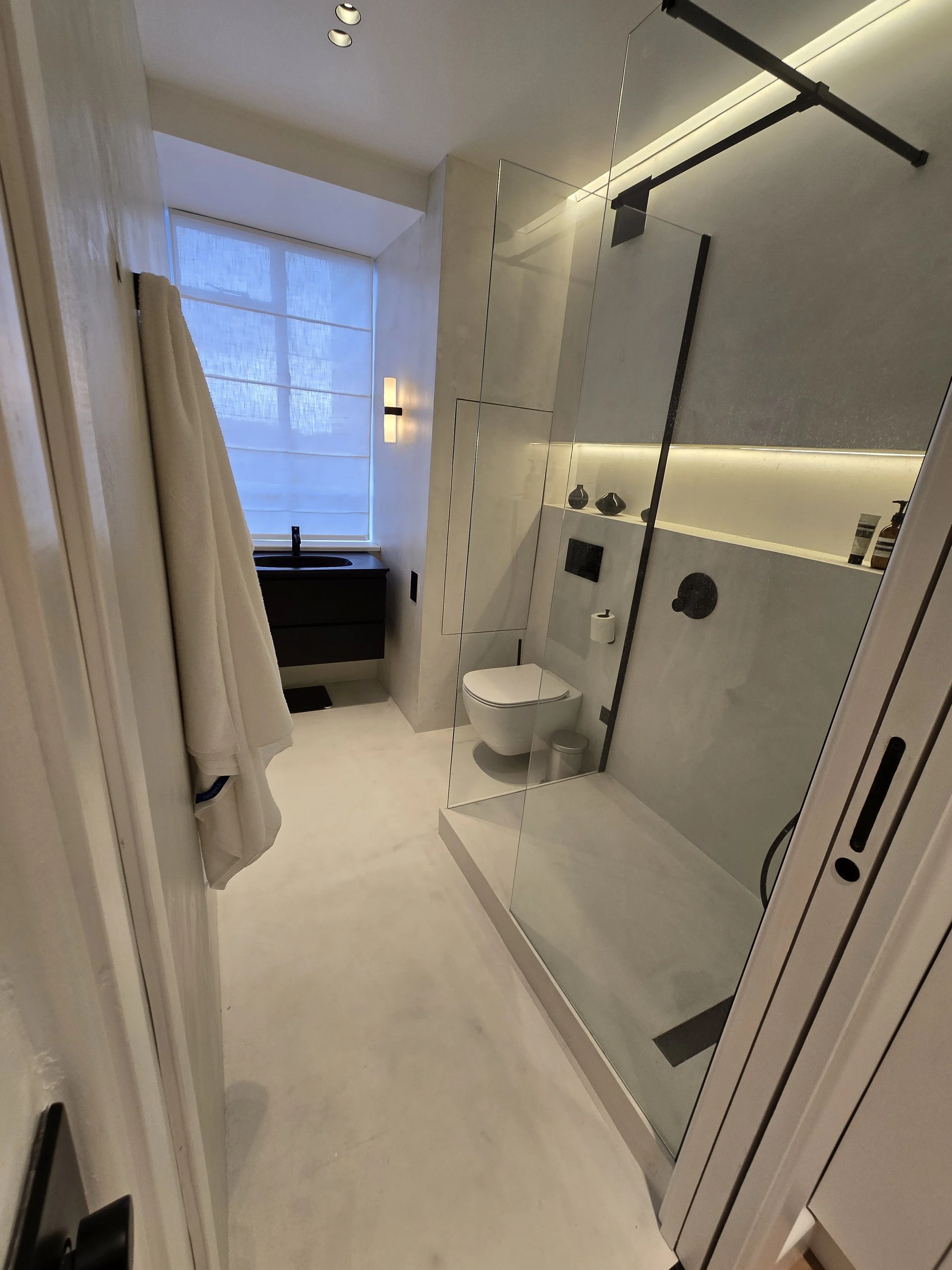 Modern bathroom with a glass-enclosed shower, a wall-mounted toilet, a black vanity with a sink, and a large window with a curtain, illuminated by natural light and soft interior lighting.