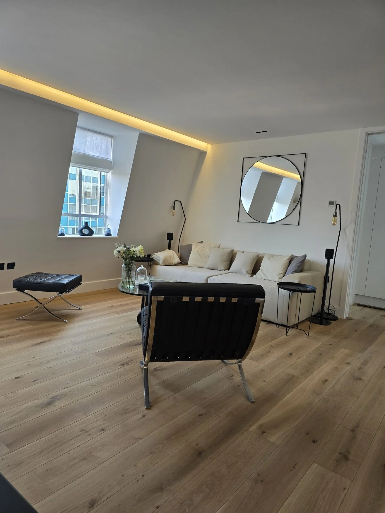 Modern living room with a white couch, black accent chairs, a round mirror on the wall, a window with a city view, and wood flooring.