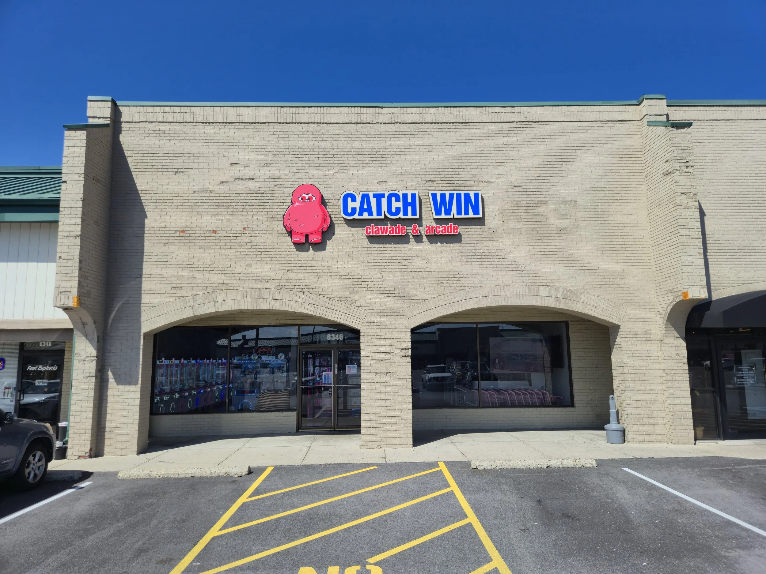The front of a brick building with a sign that reads 'Catch Win Clawade & Arcade' with a pink mascot. The building has large windows, and there are parking spaces and yellow striped lines marked on the asphalt in front.