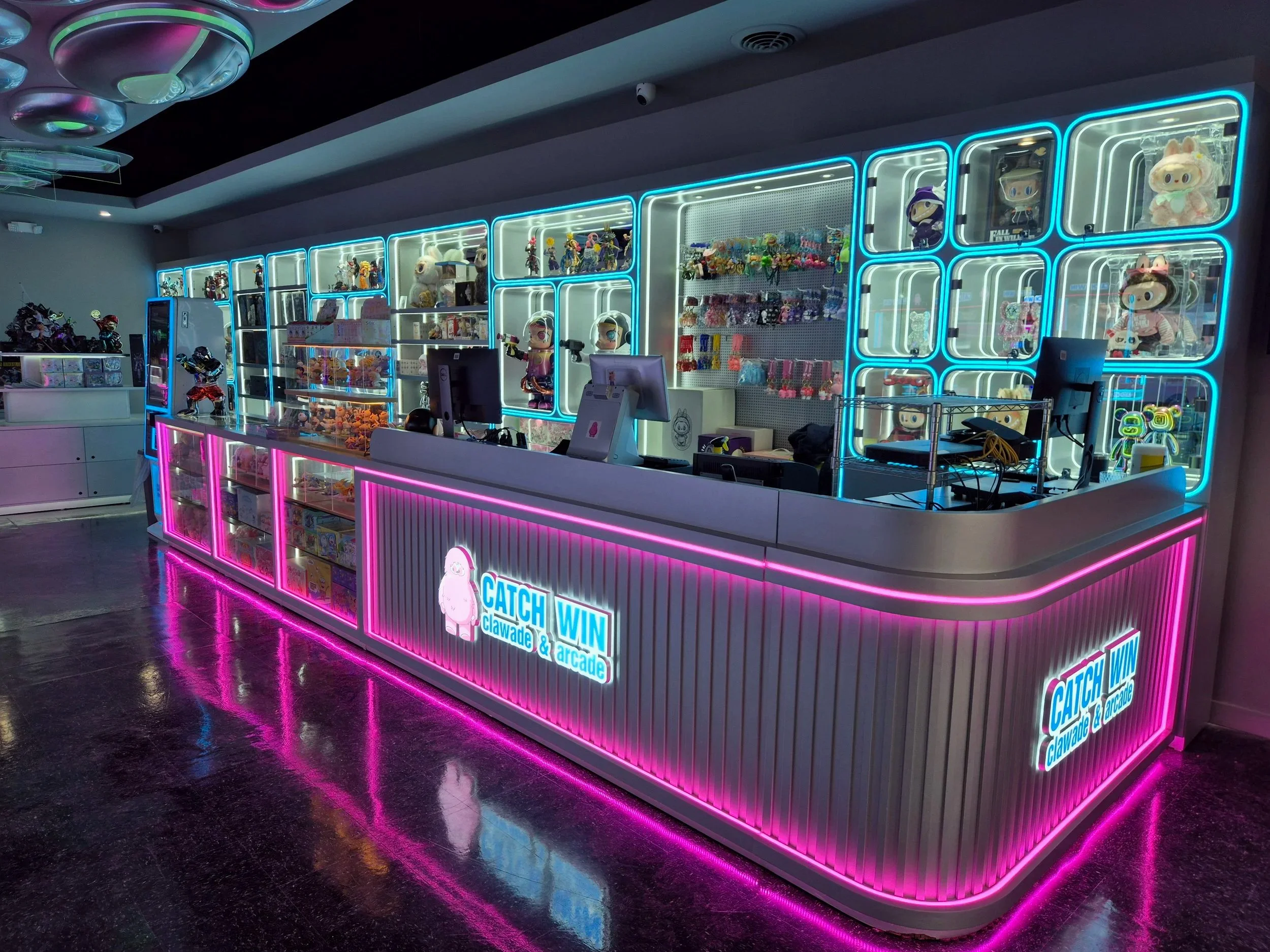 A brightly lit arcade counter with neon pink, blue, and white lights. The counter displays plush toys and figurines, and signs that read 'Catch, Claw & Win' and 'Claw Arcade.' The background features illuminated shelves with more toys and prizes.