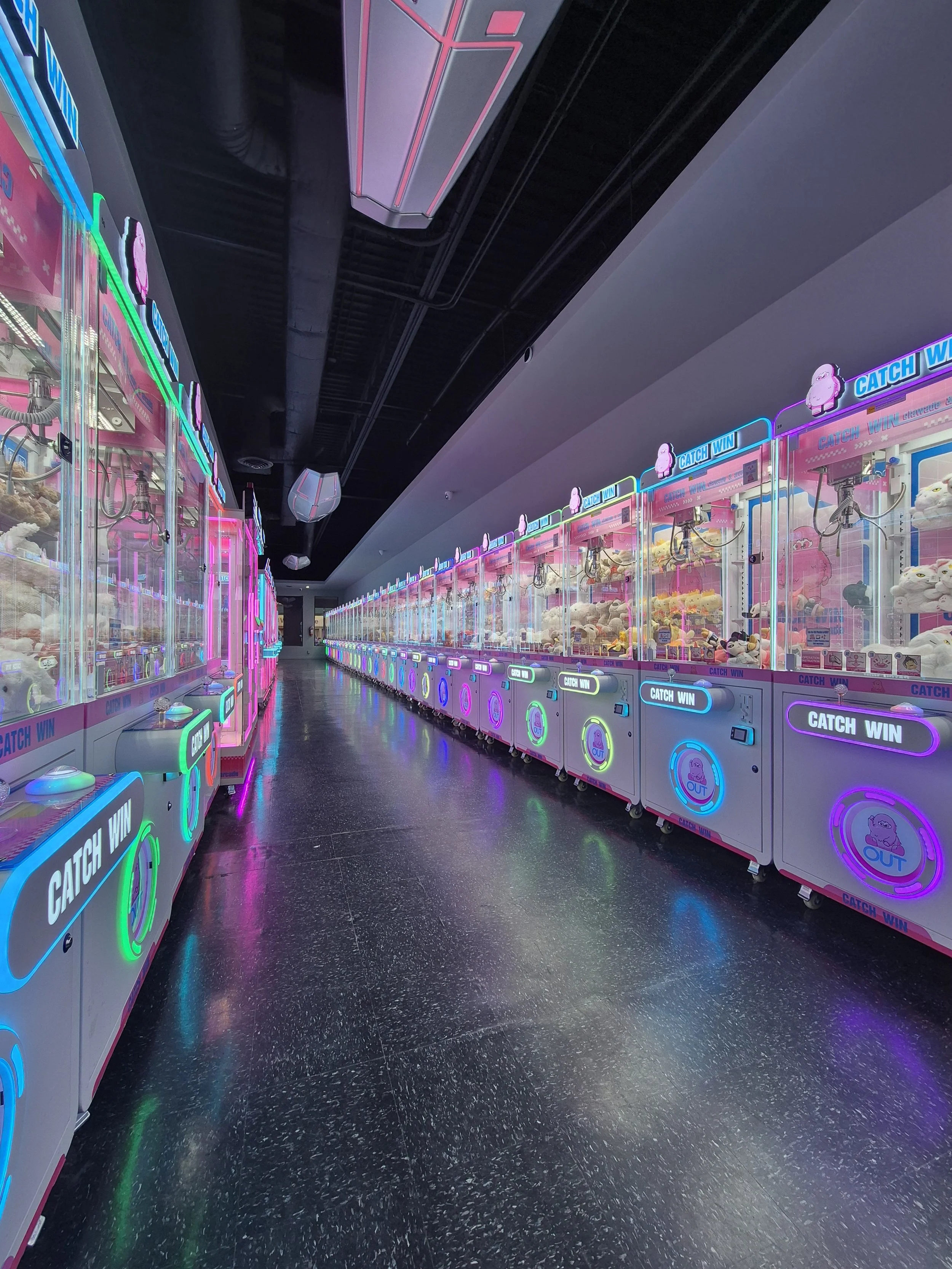 Row of colorful claw machines filled with plush toys inside an arcade with neon signs and a black shiny floor.