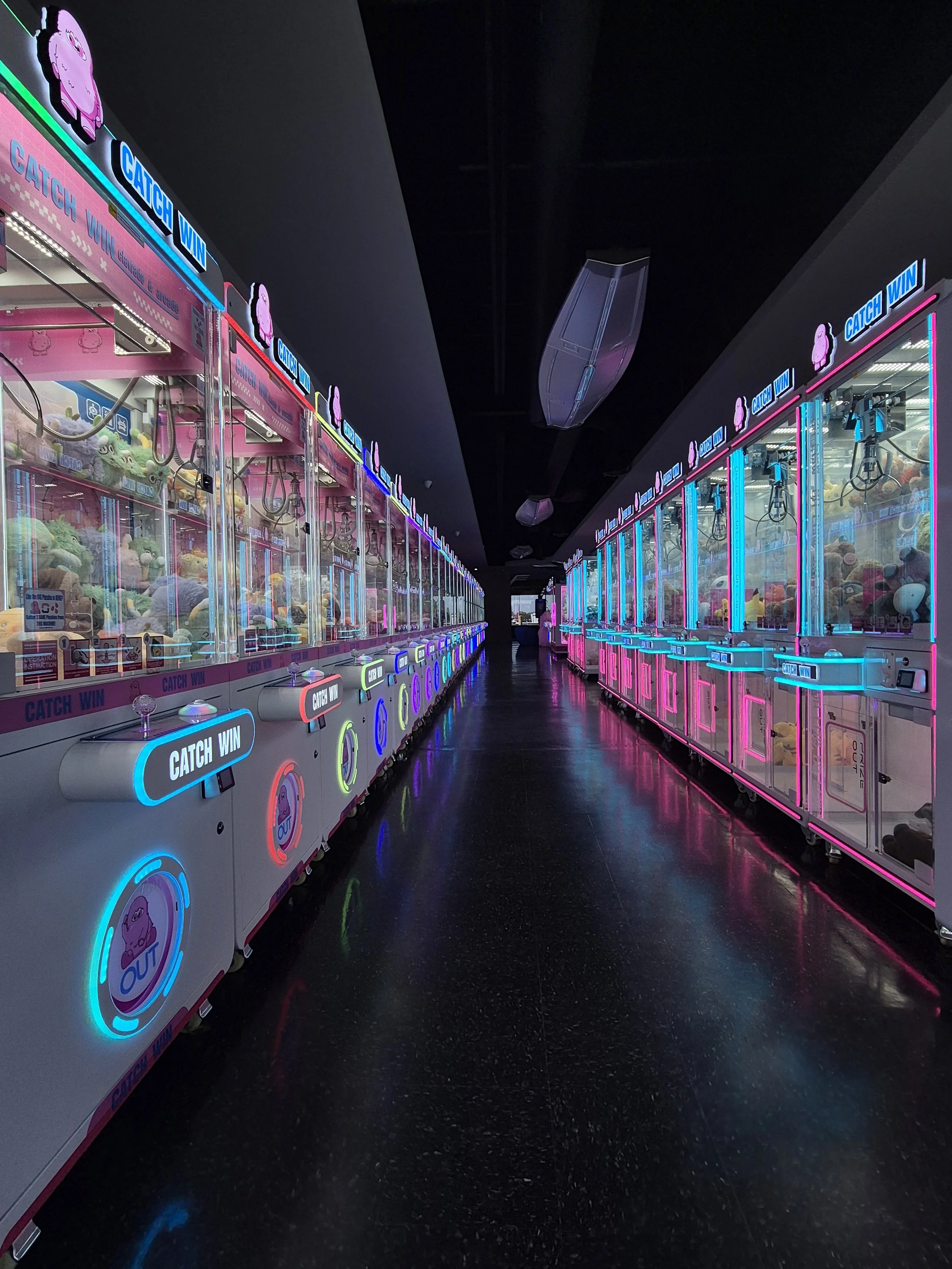 Rows of pink and white claw machines with plush toys, illuminated by neon pink, blue, and yellow lights, in an arcade setting.