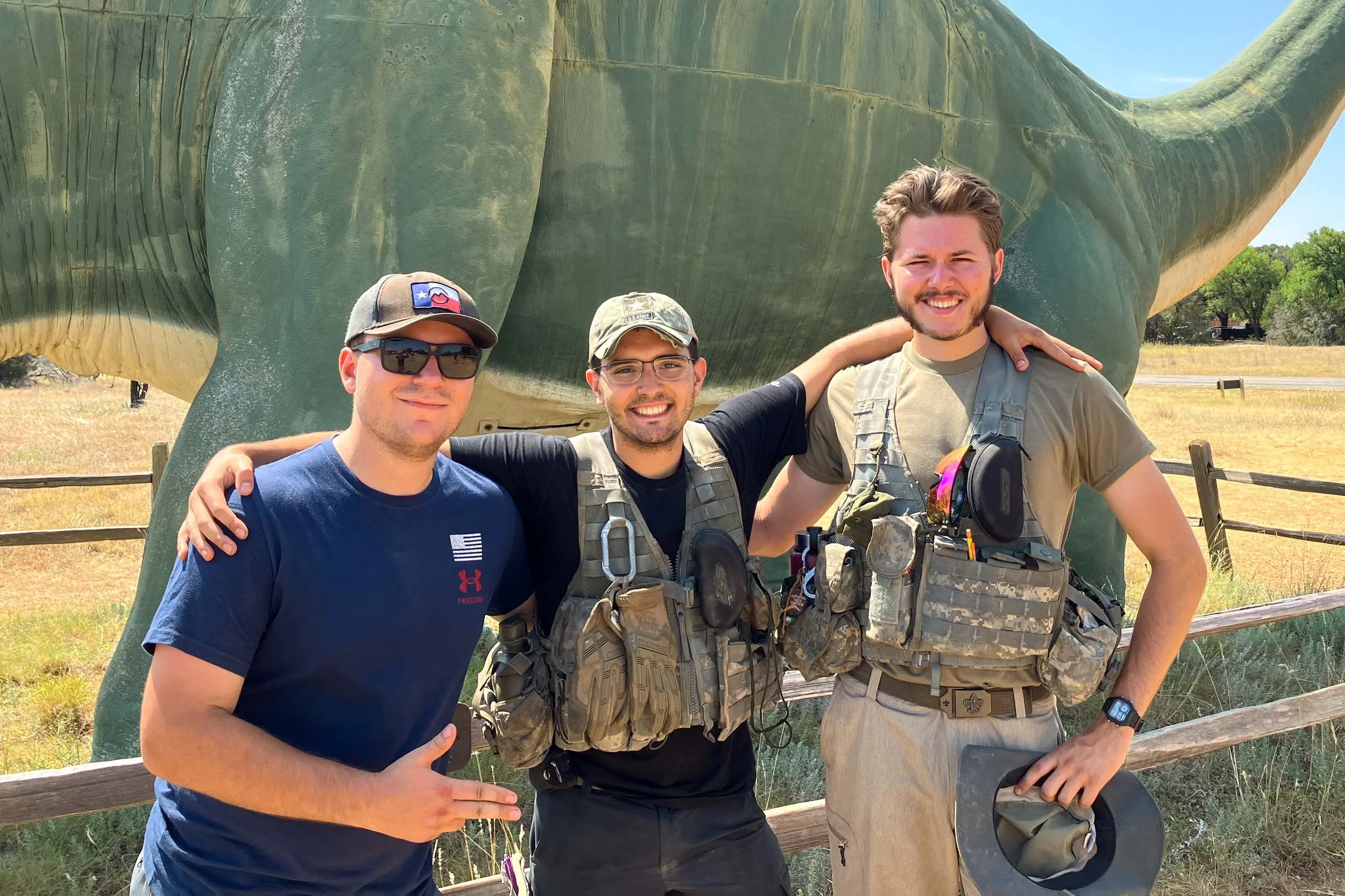 Three men standing together outdoors in front of a large green dinosaur statue, smiling and posing with their arms around each other, during daytime with clear sky.