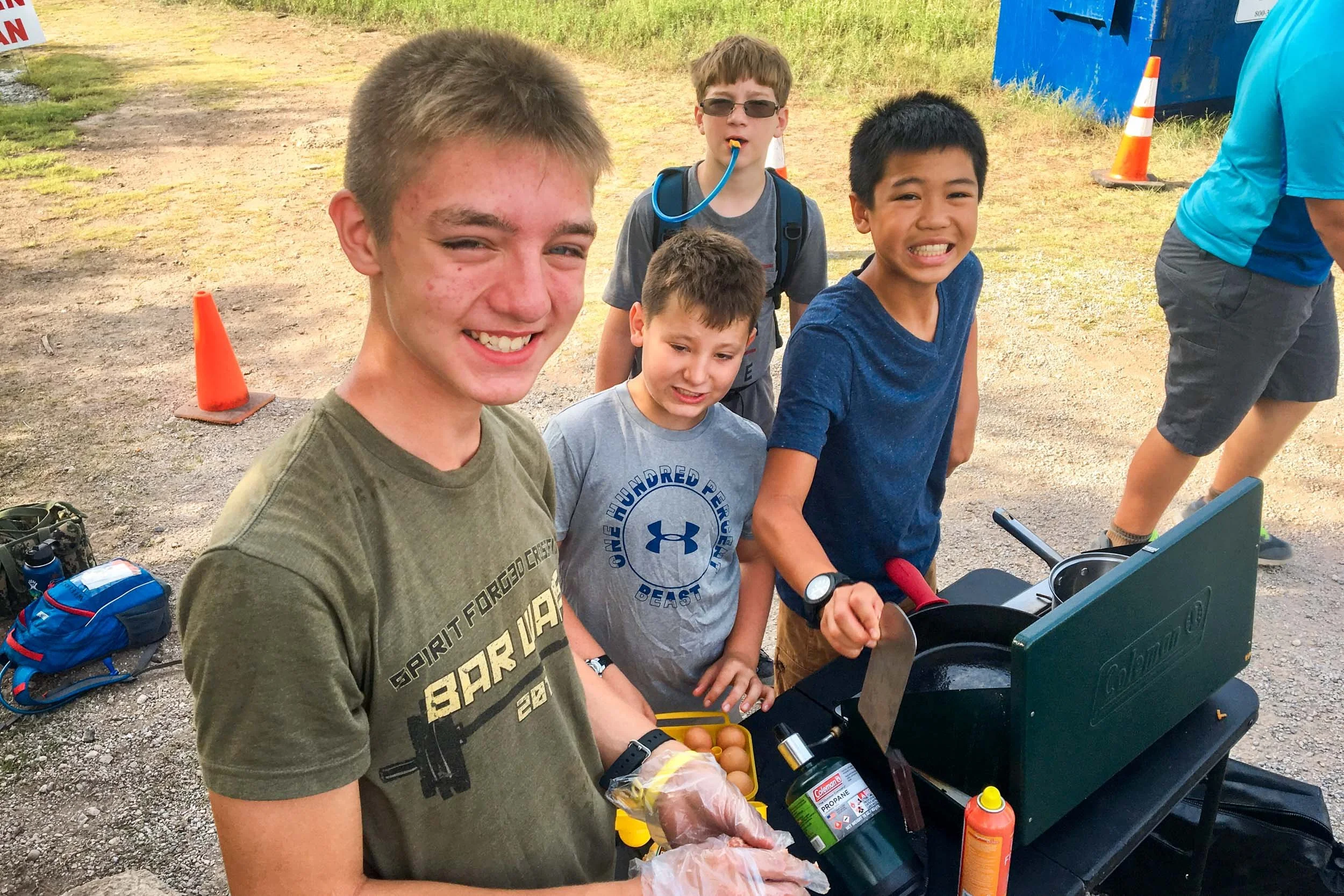 A group of boys gathered around a outdoor grill cooking breakfast.