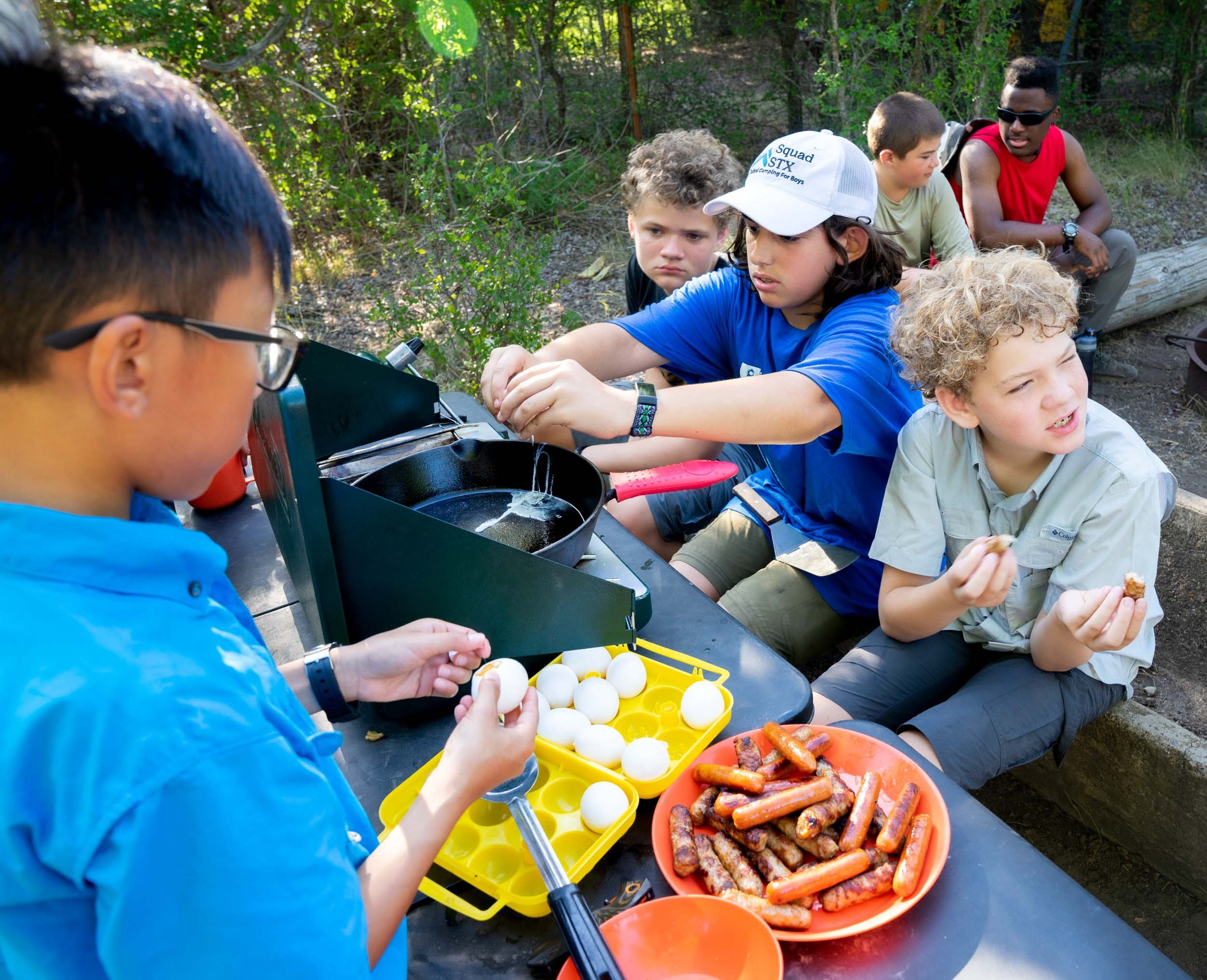 Campers preparing breakfast food outdoors, including eggs and sausages.