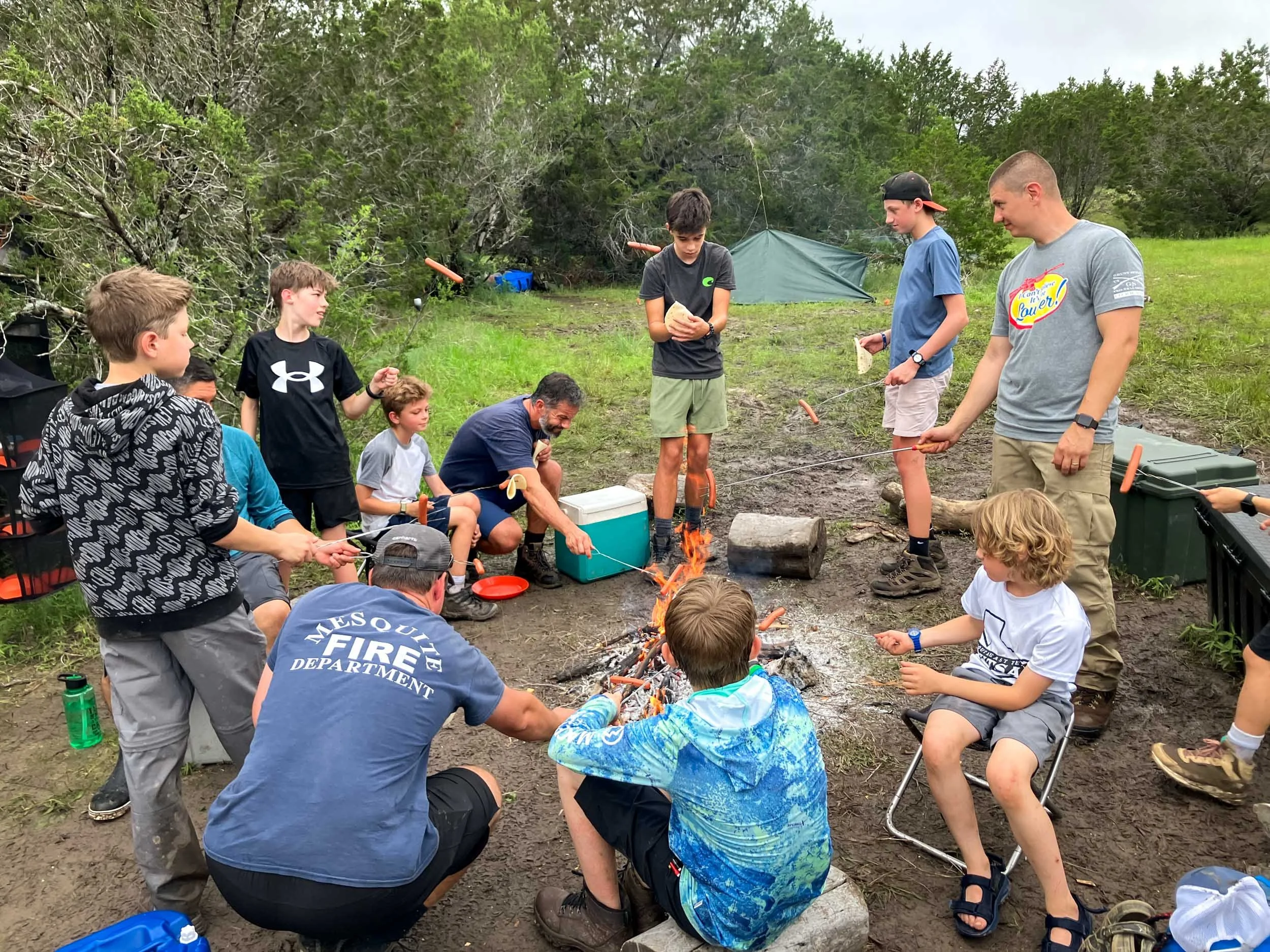 Group of children and teenagers gathered around a small outdoor campfire, some roasting hot dogs on sticks, in a wooded area with a tent in the background.