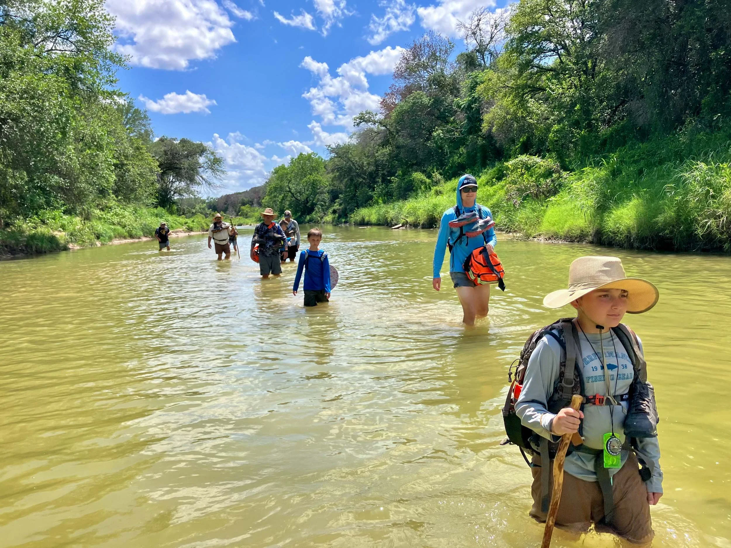 Fathers and sons hiking through a shallow river on a sunny day.