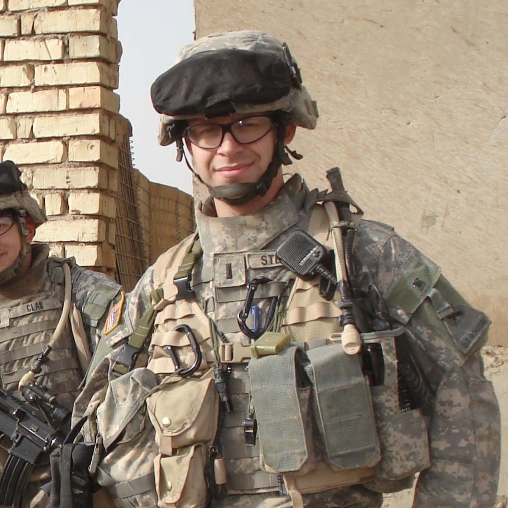 A soldier wearing camouflage uniform, tactical gear, helmet, glasses, and winking, standing outdoors against a brick wall.