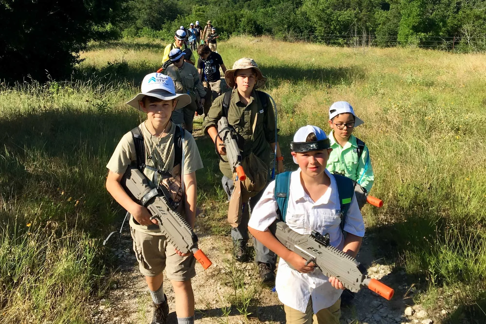 Group of boys walking in a line through a grassy outdoor area, carrying laser tag guns.