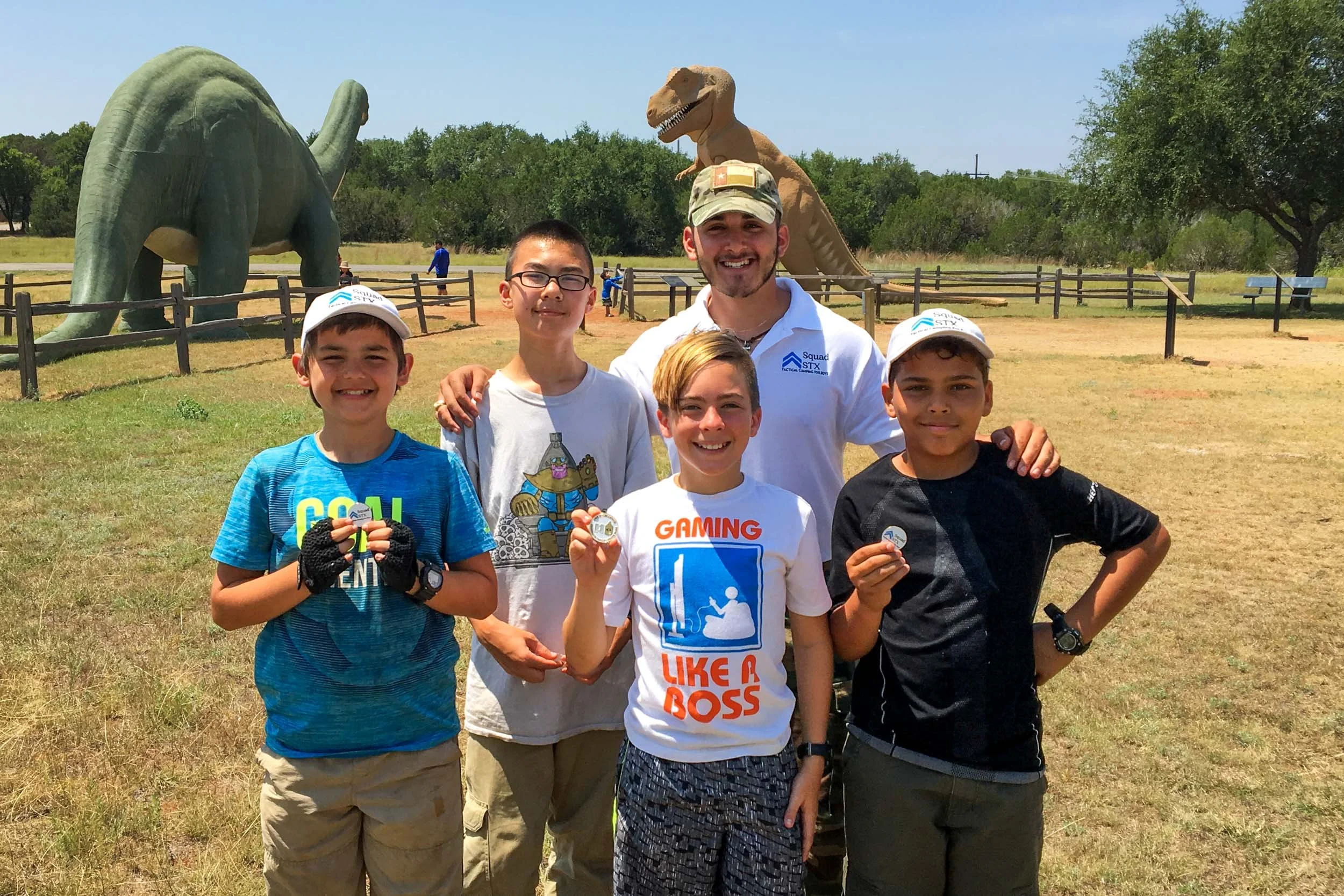 Group of five people, including four children and an adult, smiling outdoors in front of large dinosaur statues at a park.