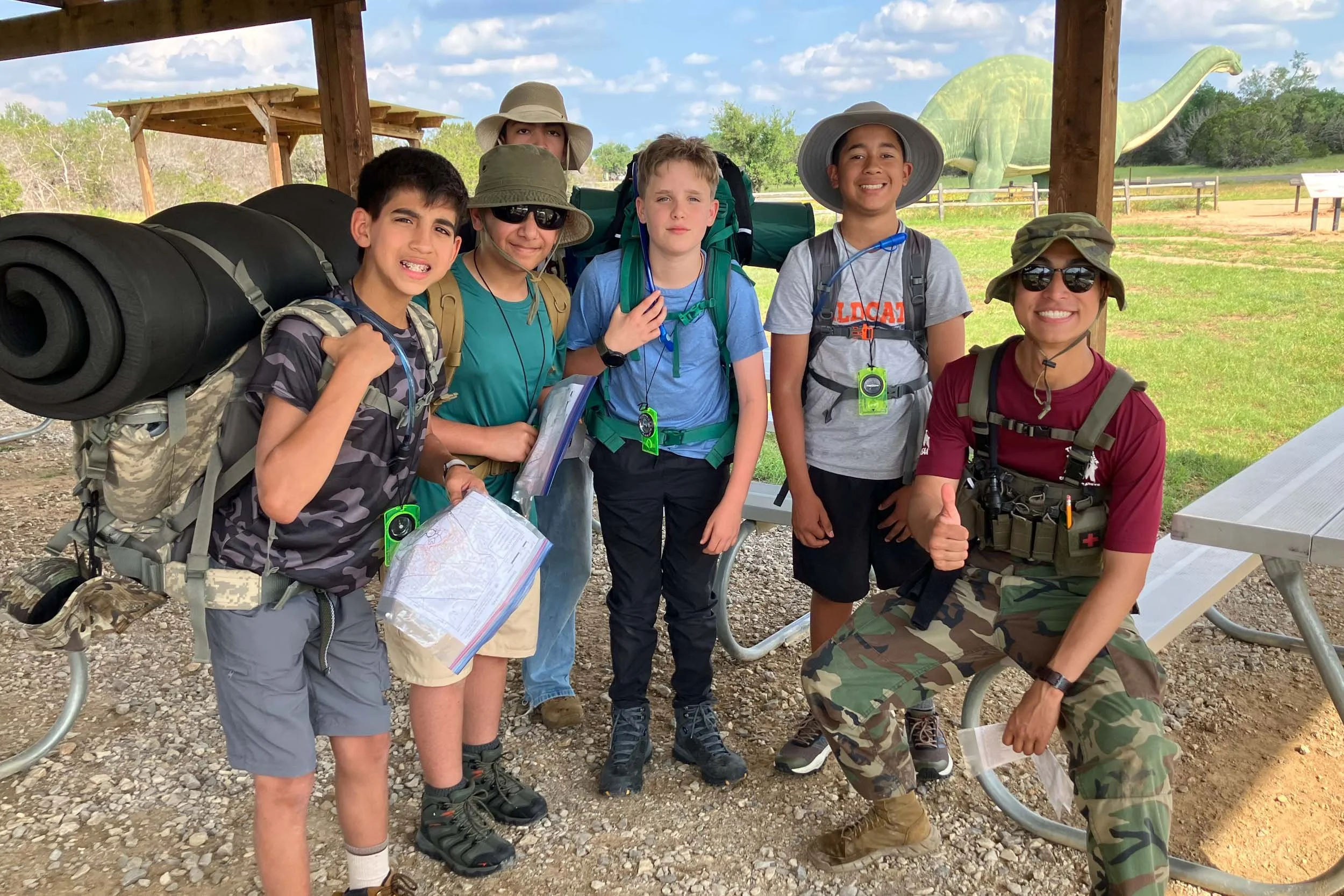 Group of six children and one adult woman posing outdoors with a dinosaur statue in the background. They are dressed for outdoor activity, wearing hats, backpacks, and casual clothing, and are smiling.
