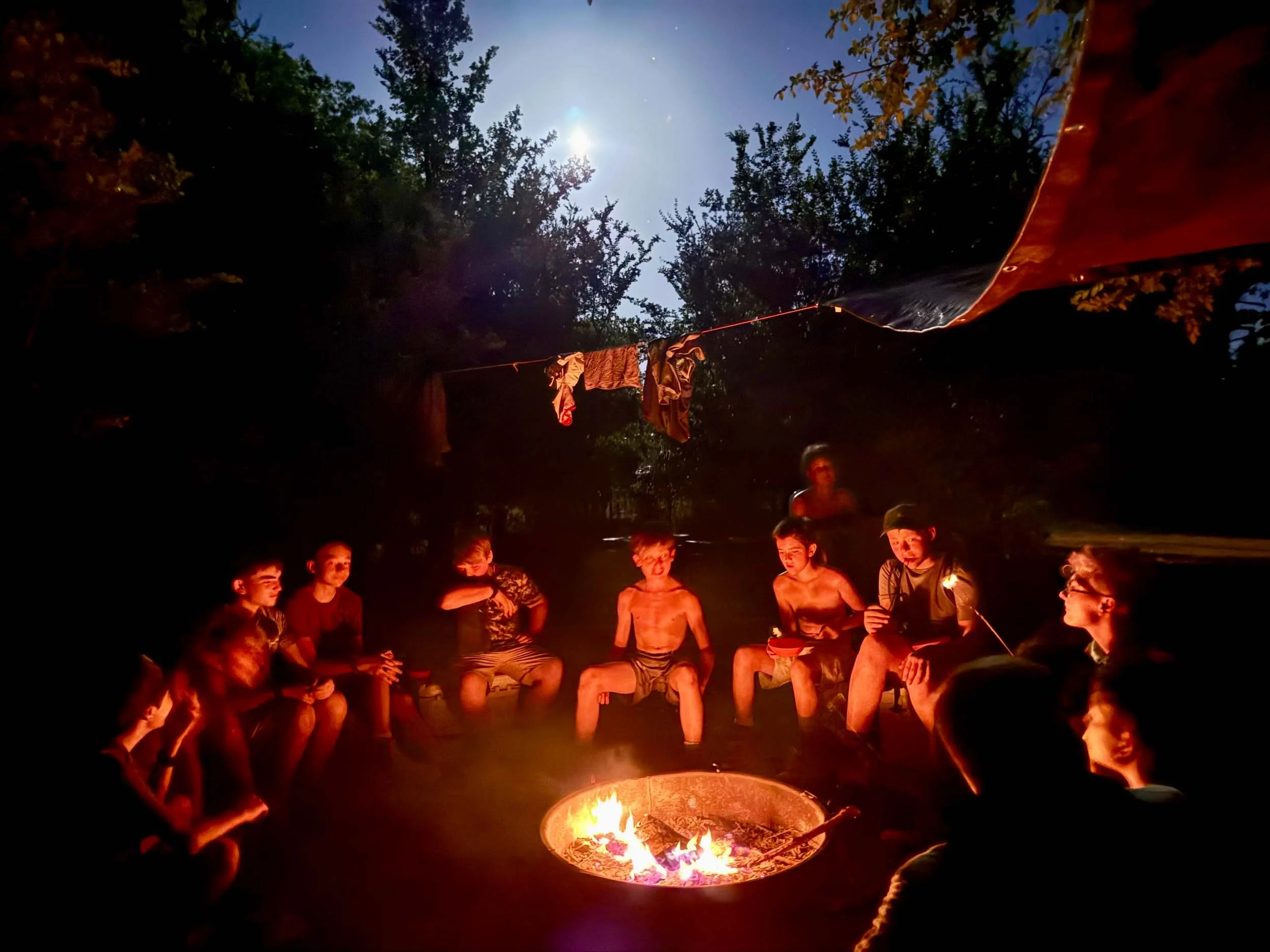 Boys gathered around a campfire at night with a moonlight sky behind them.