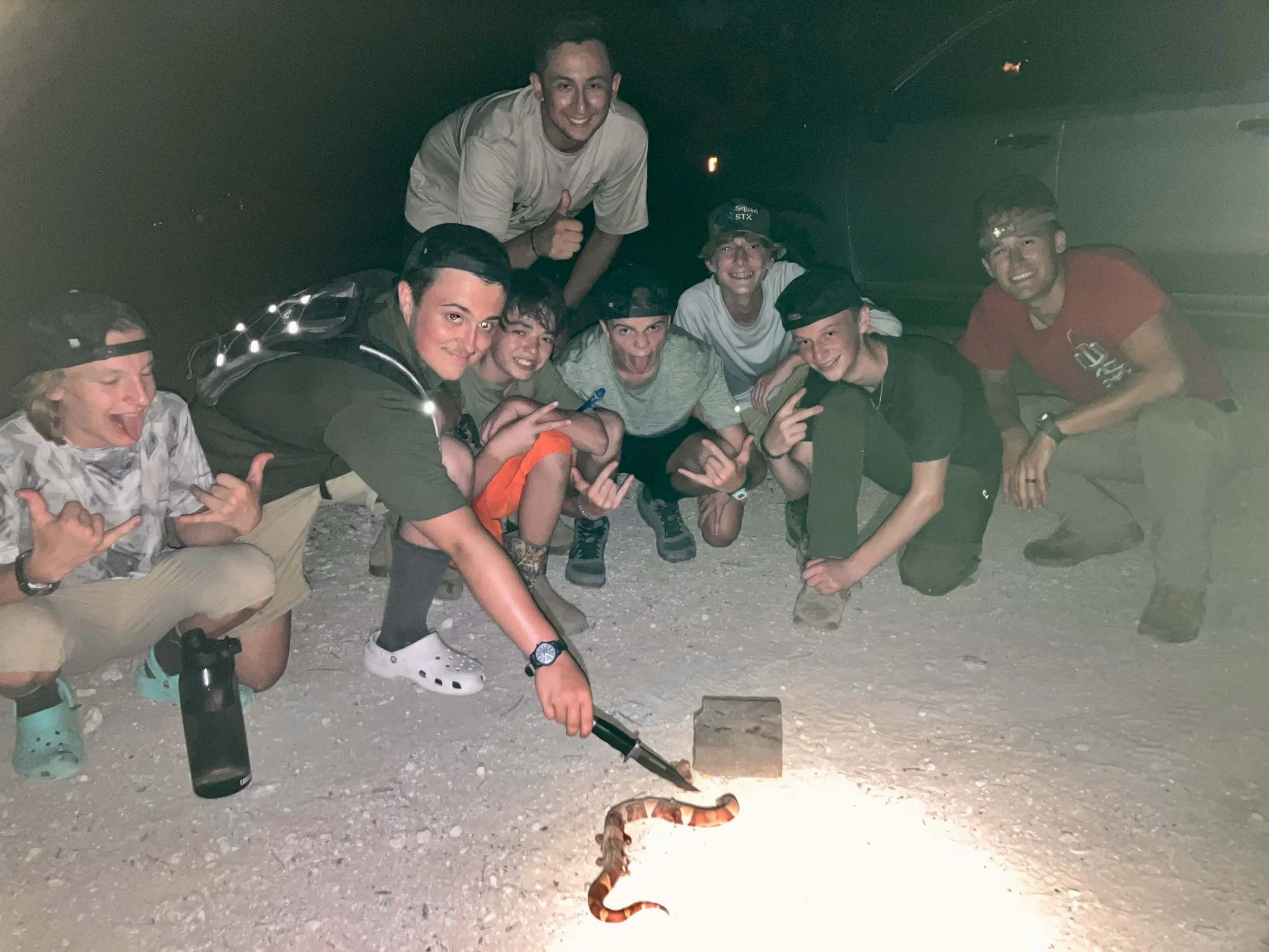 Group of young men crouching and smiling at night around a small fire with a snake on the ground in front of them.