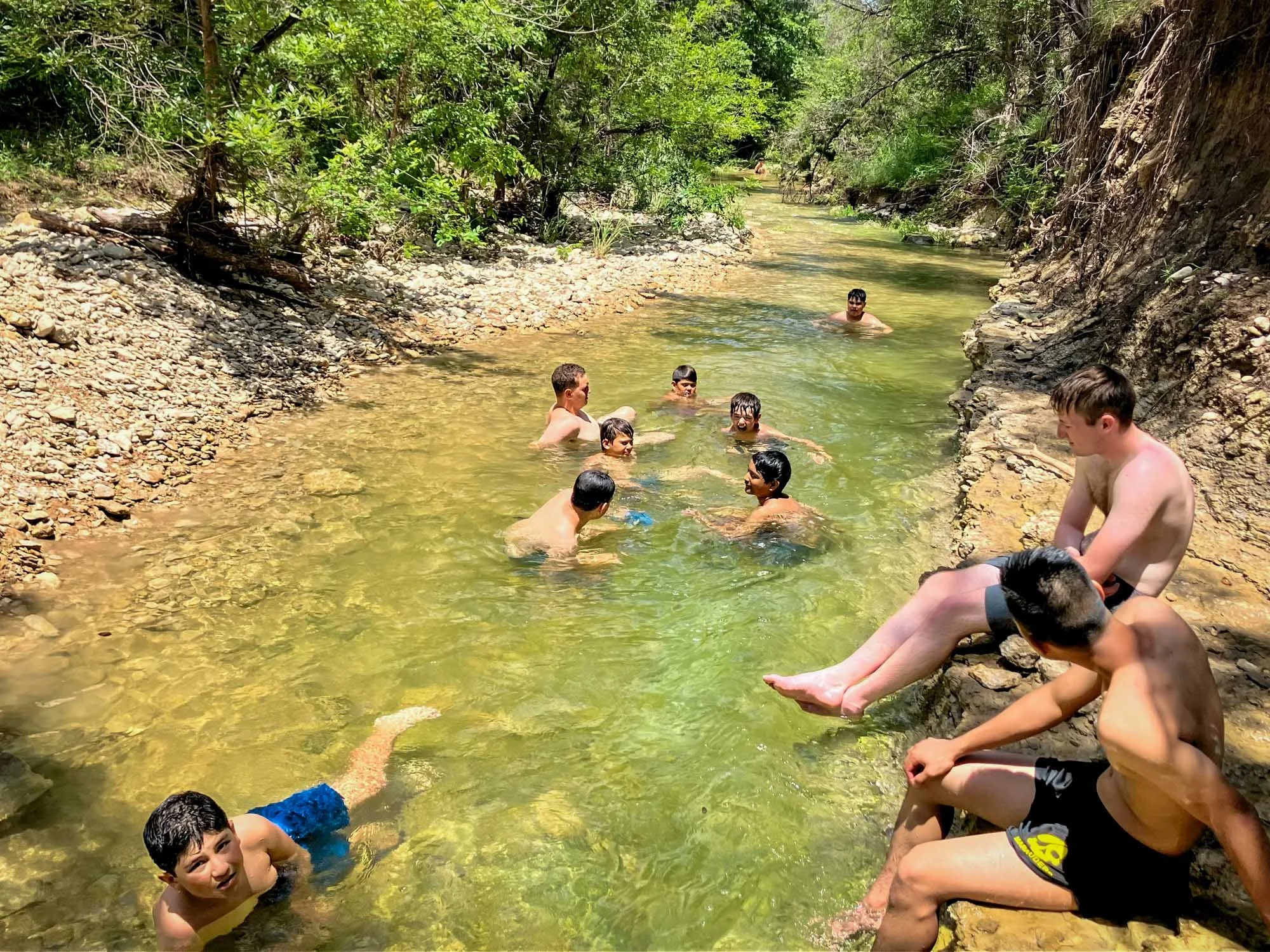 Campers swimming and relaxing in a shallow creek surrounded by nature.
