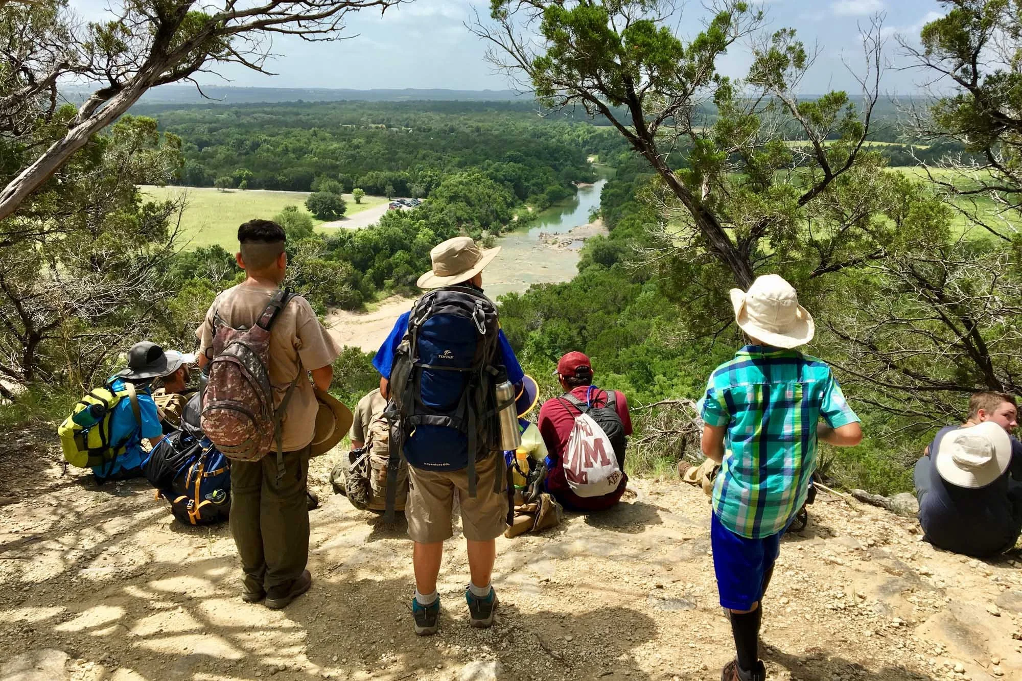 Group of hikers sitting and standing on a hill overlooking a river and lush green forested landscape under a cloudy sky.