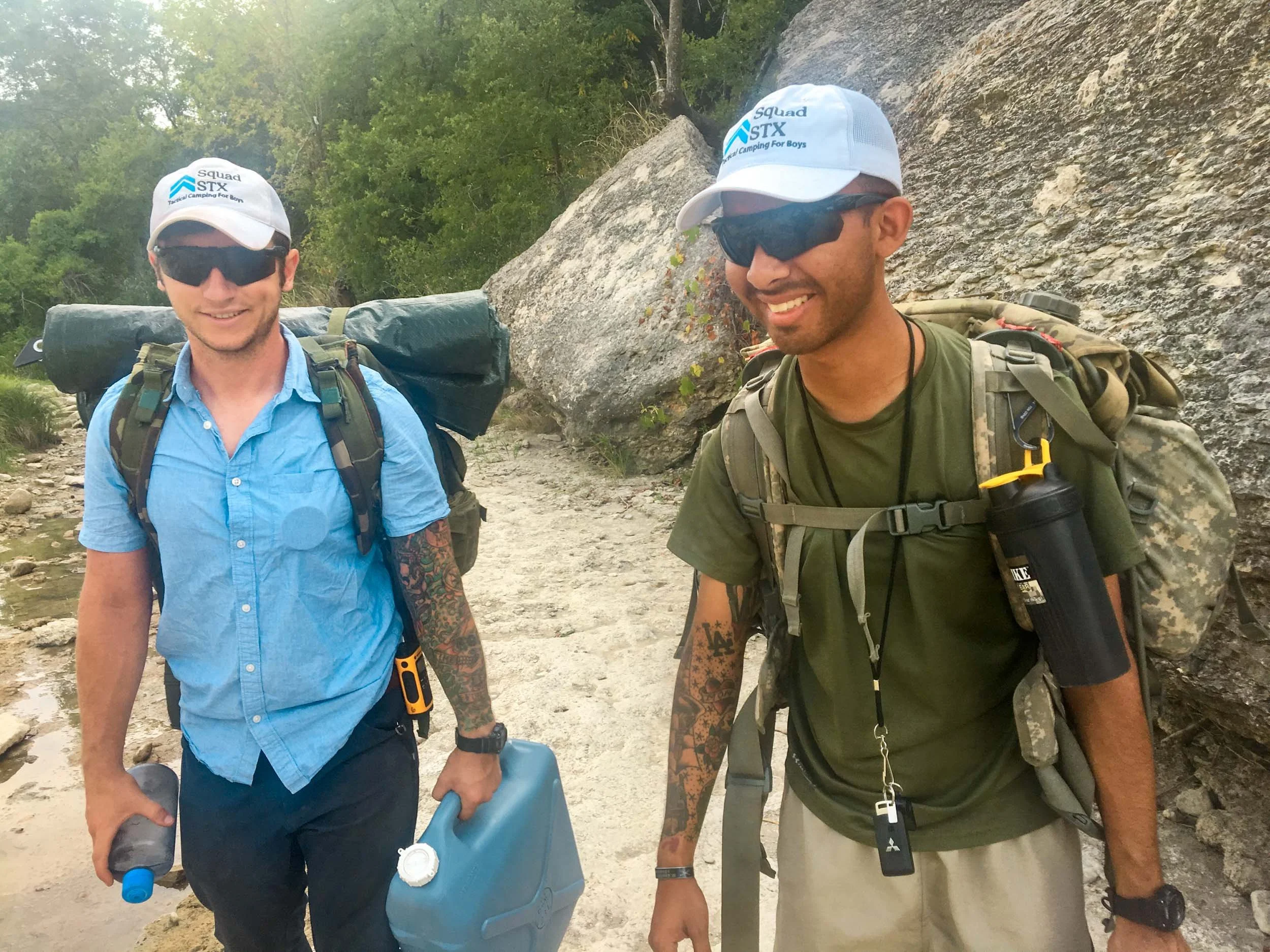 Two men geared up for outdoor hiking, standing beside a rocky area with trees and a creek, wearing baseball caps, sunglasses, and backpacks.