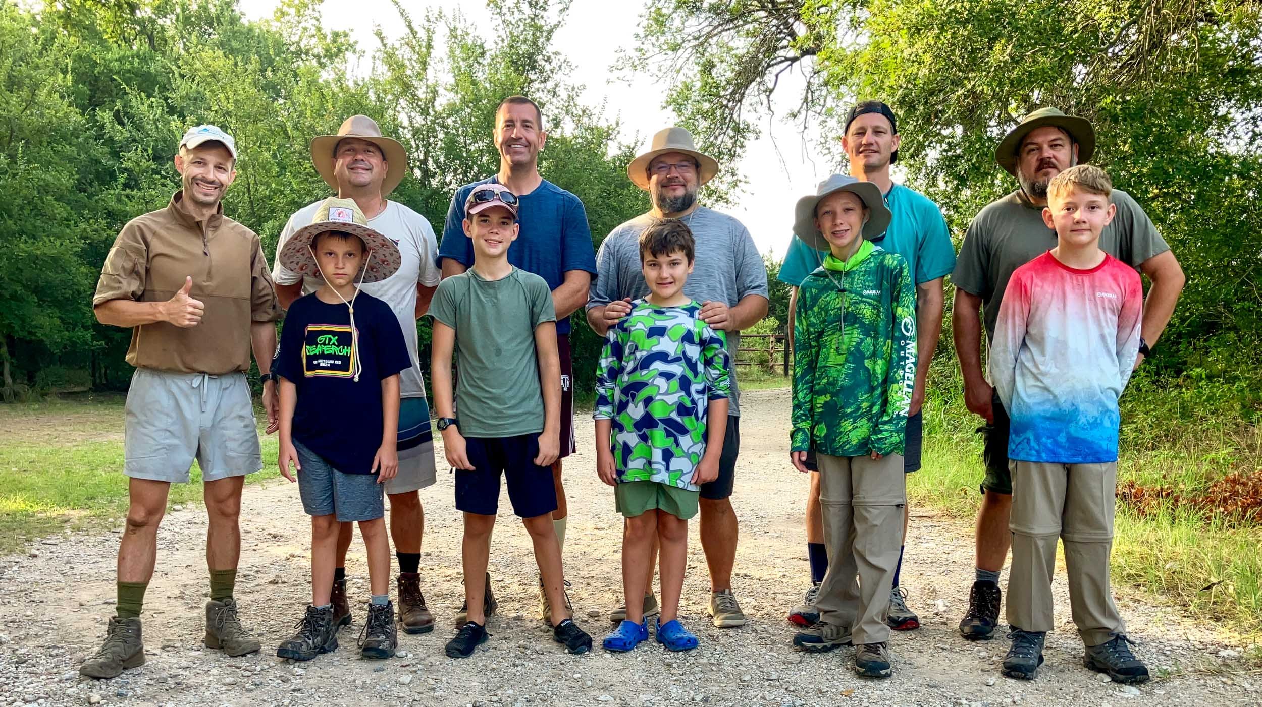 Group of ten people, including children and adults, standing outdoors on a sunny day with trees in the background. Most are dressed in casual outdoor clothing and hats, some smiling and one giving a thumbs up.