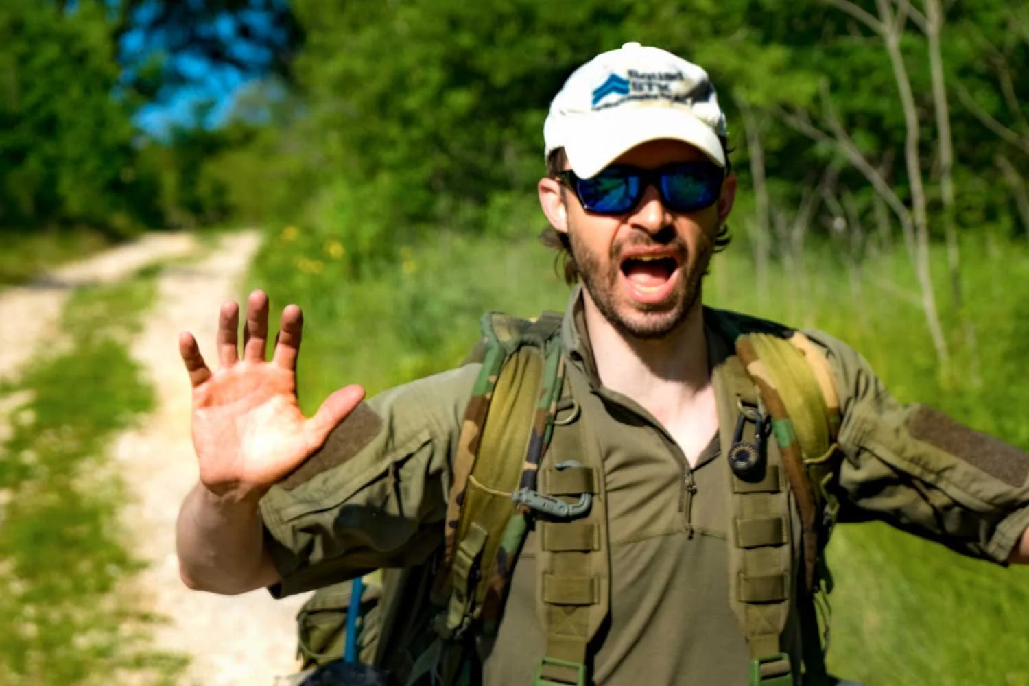 A man in camouflage clothing, wearing sunglasses, a white cap, and a backpack, reaches out with his left hand while standing on a dirt path surrounded by green trees.