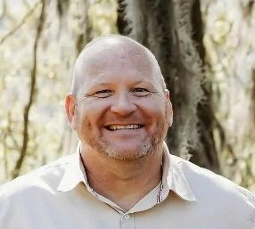 A smiling man with a bald head and beard, wearing a light-colored collared shirt, standing outdoors with trees in the background.
