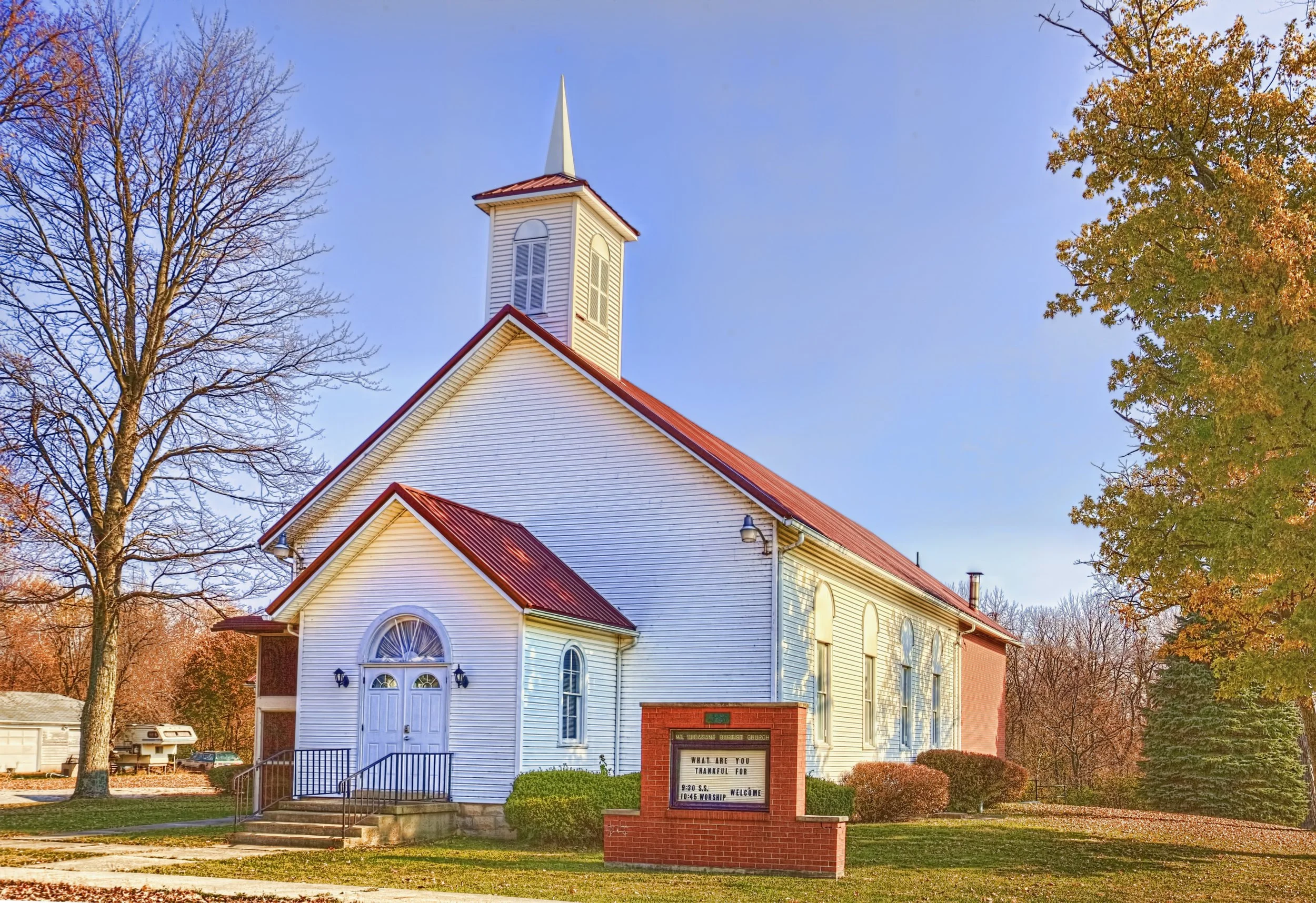 A white church with a tall steeple, surrounded by autumn trees with colorful leaves, and a sign in front displaying church service times.