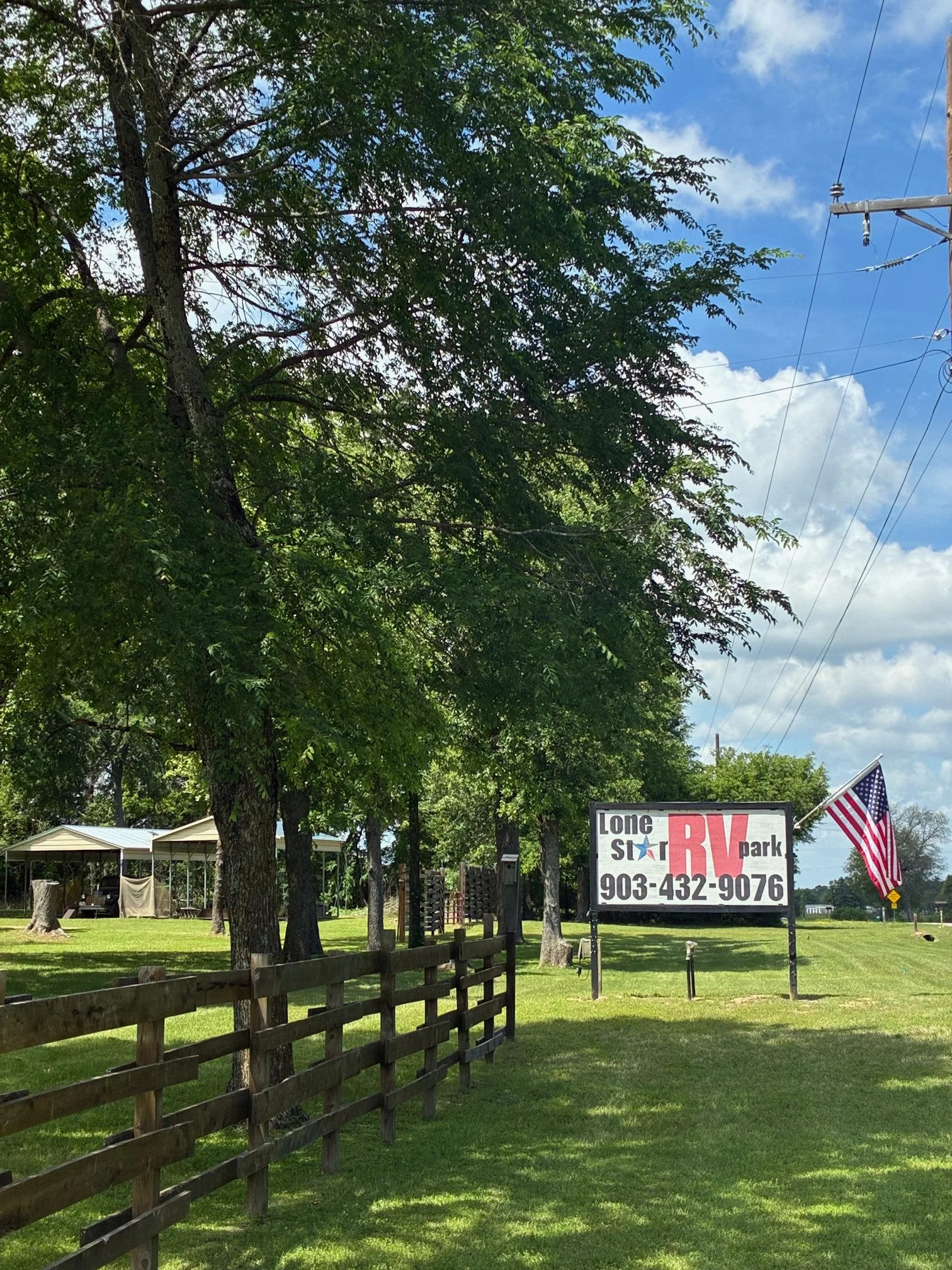 Sign for Lone Star RV Park with American flag, green grassy area, trees, blue sky with some clouds.
