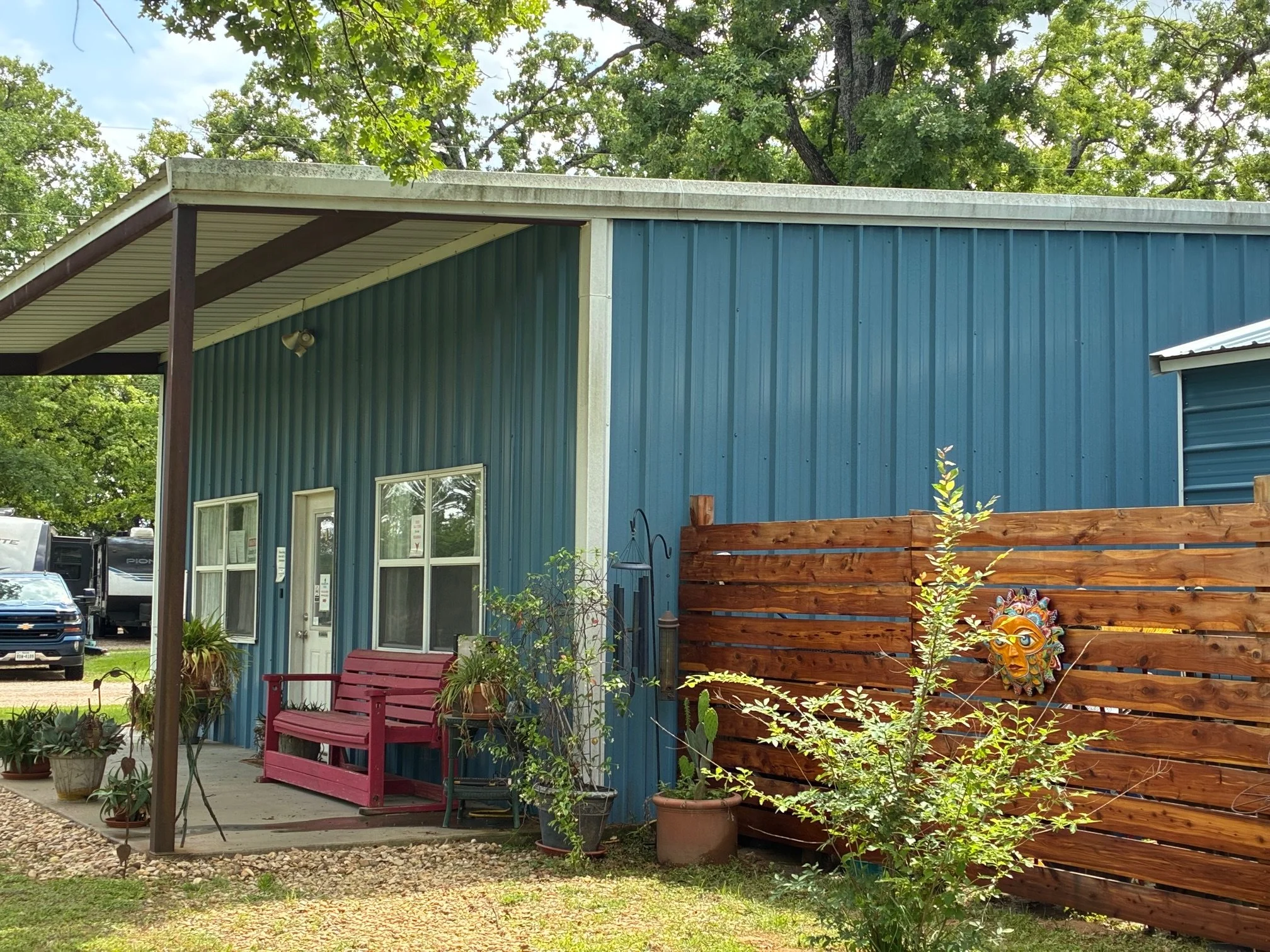 Front of a blue metal building with a red porch bench, potted plants, and a decorative sun mask on a wooden fence.