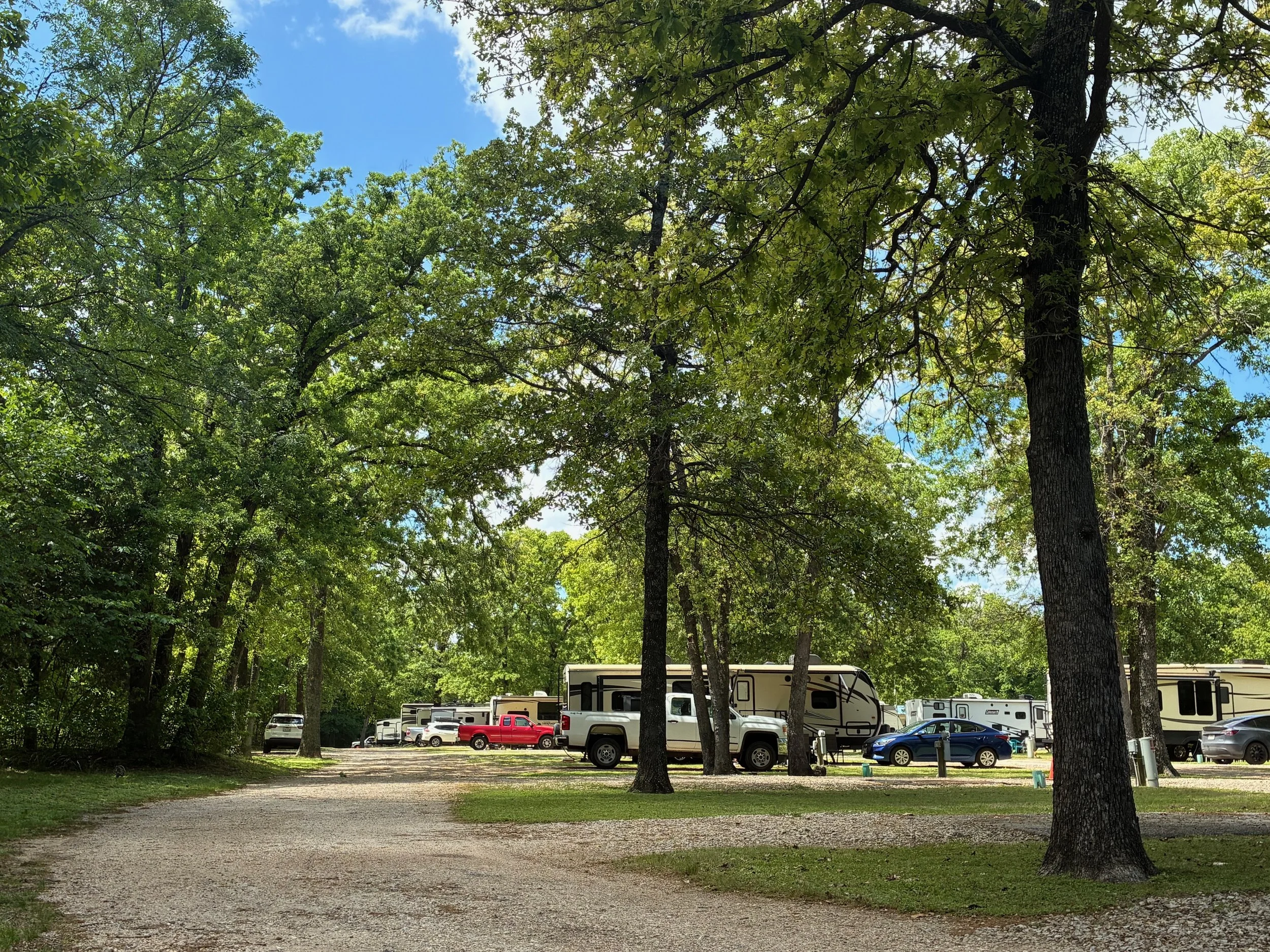 A campground with trees, several campers, trucks, and cars parked on gravel paths under a bright, partly cloudy sky.