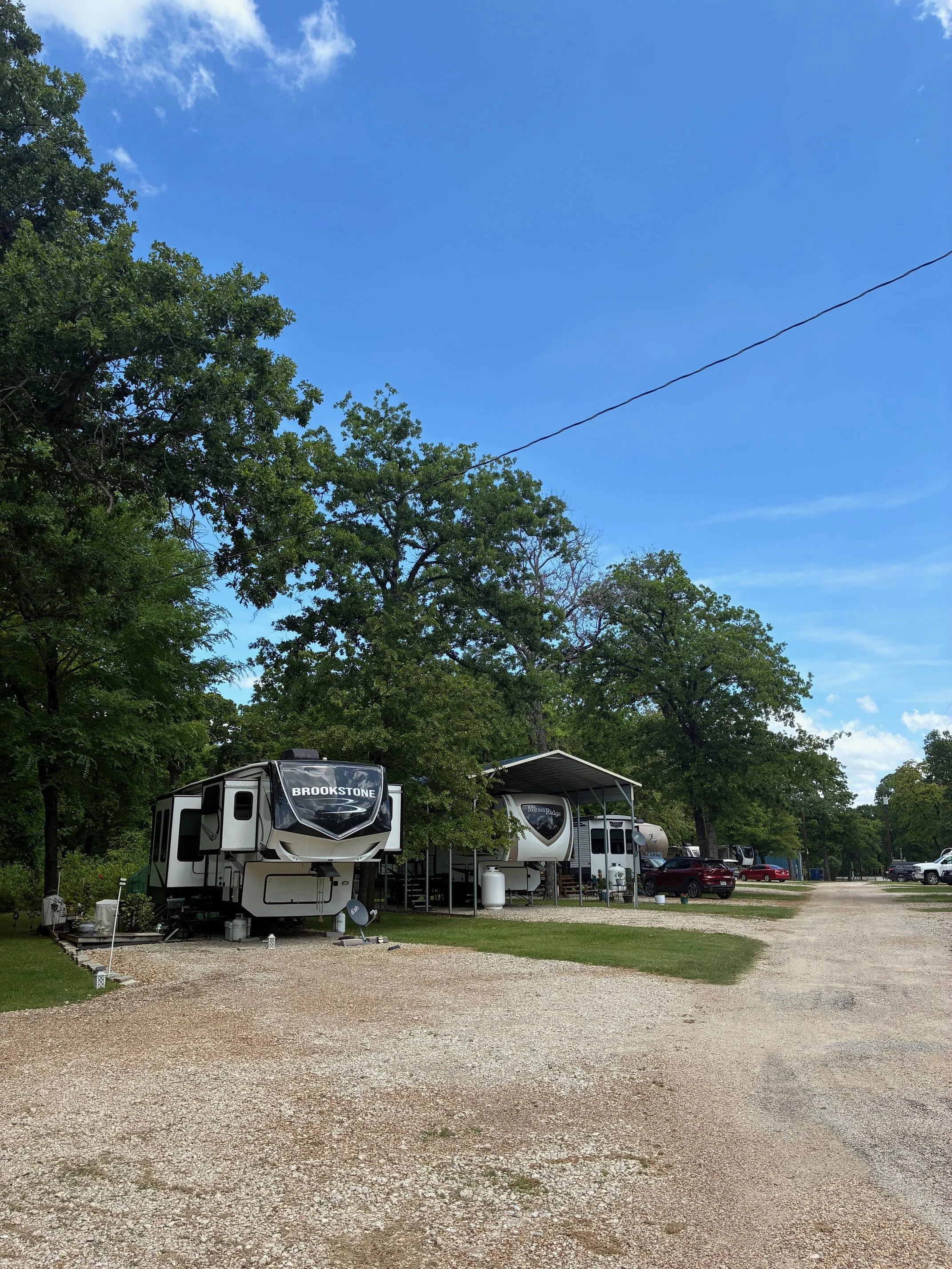 A campground with several RVs and cars parked on a gravel lot, surrounded by green trees and a blue sky with some clouds.