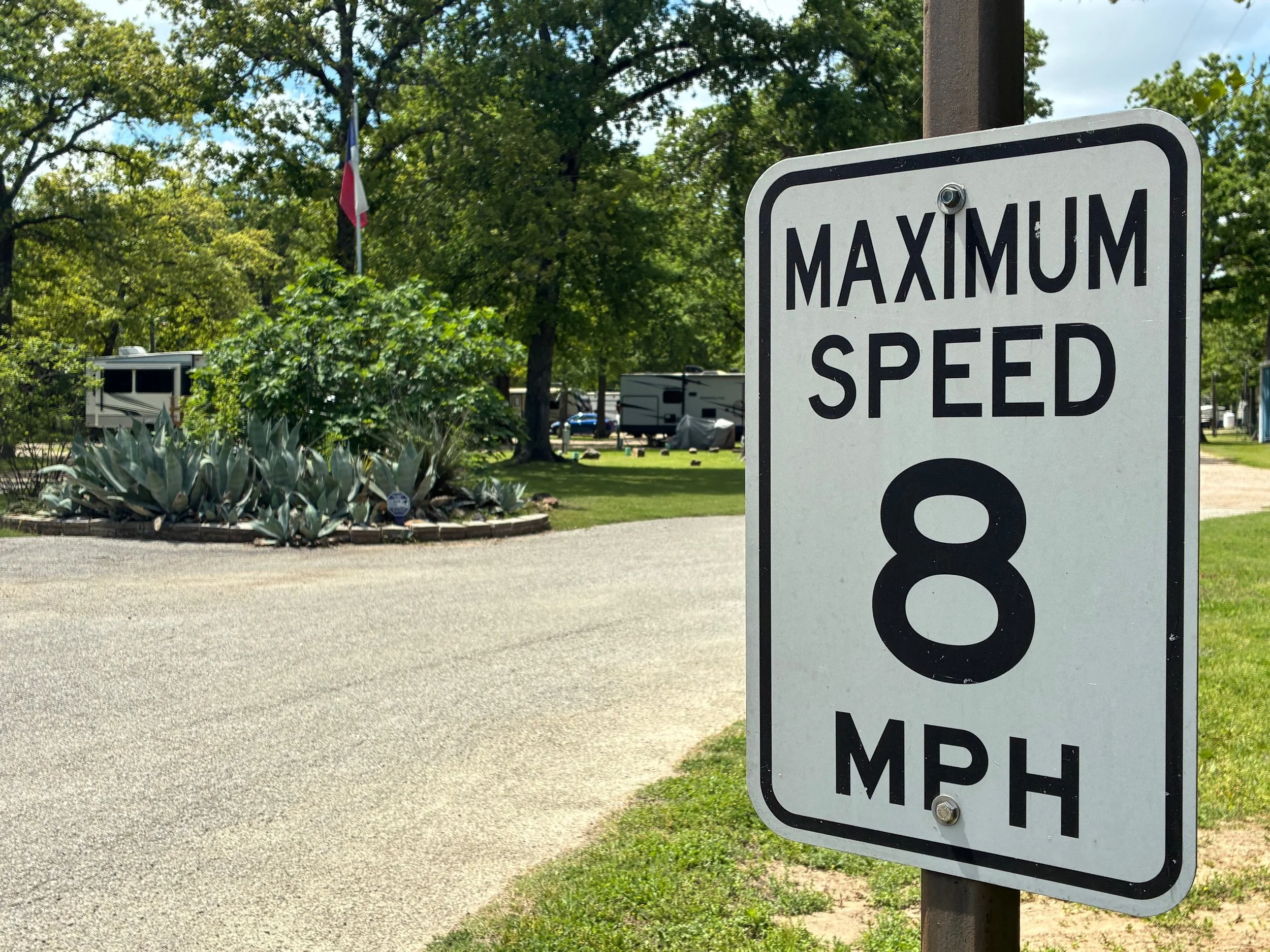 A speed limit sign stating maximum speed of 8 MPH, with a background of trees, RVs, and a flag in a park-like setting.