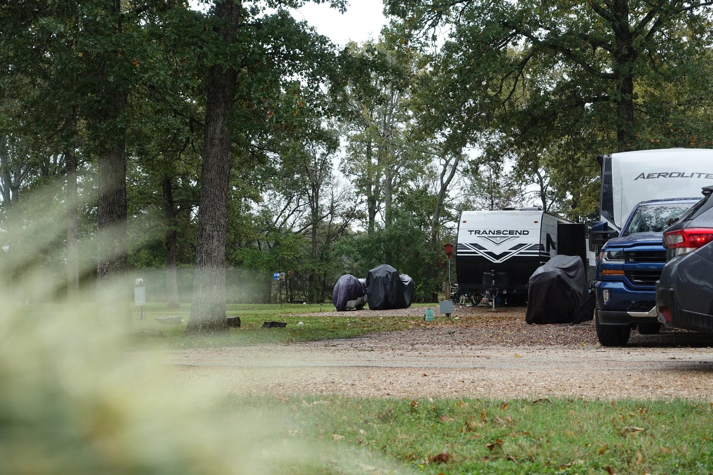 A campground with trees, RVs, and covered bikes on a dirt and grass area.
