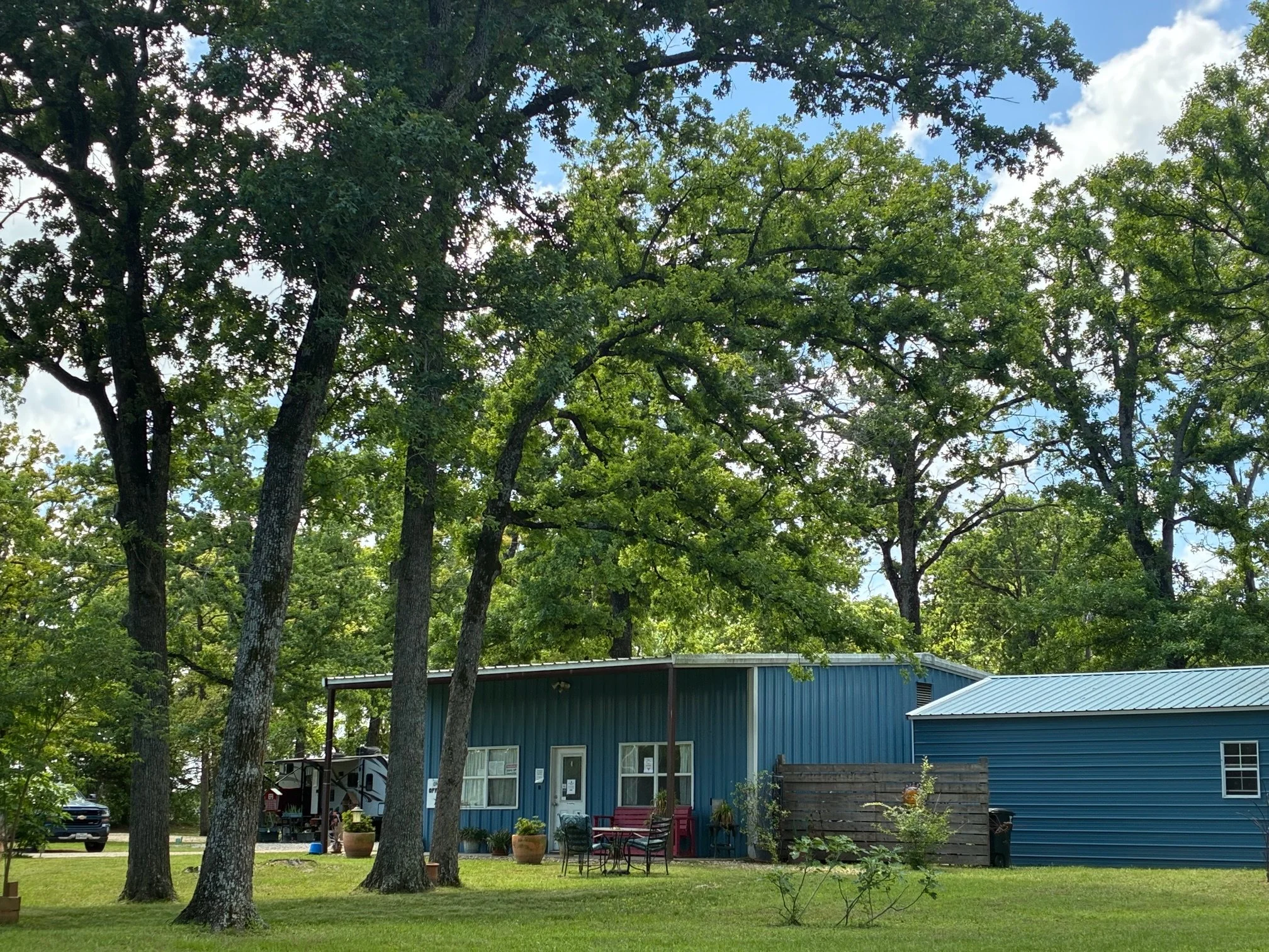 A blue metal building with a patio area and chairs, surrounded by tall green trees and grass.