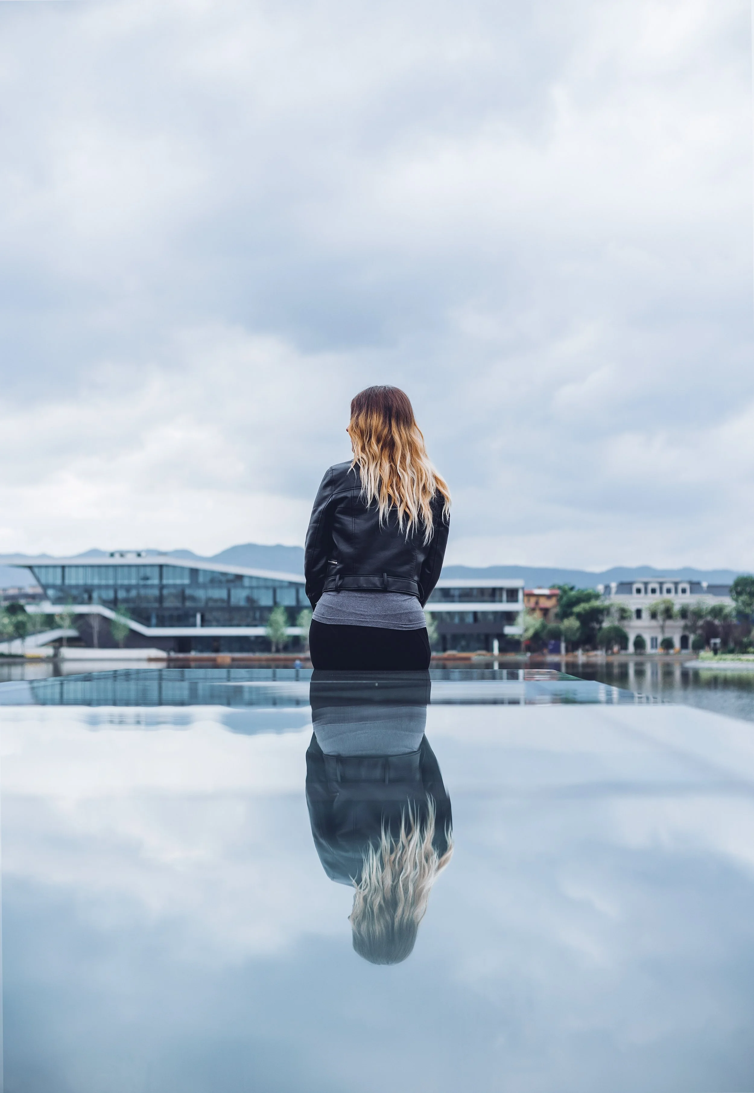 A woman with long blonde hair standing with her back facing the camera, looking over a city or townscape under a cloudy sky, with her reflection visible in a body of water in front of her.