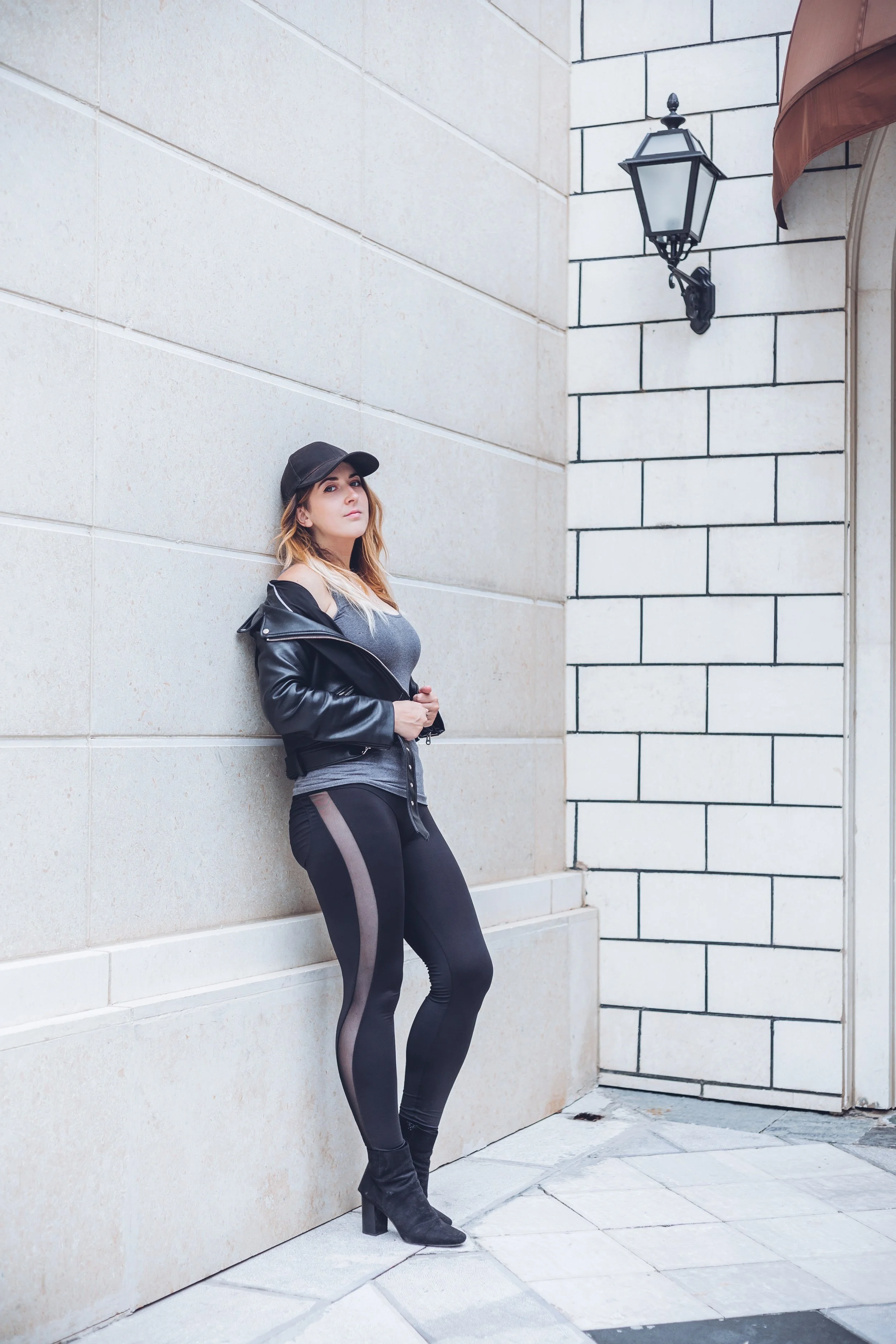 A woman with long hair wearing a black baseball cap black leather jacket gray shirt and black leggings with mesh panels, standing against a beige wall with her hand on her stomach, looking at the camera.
