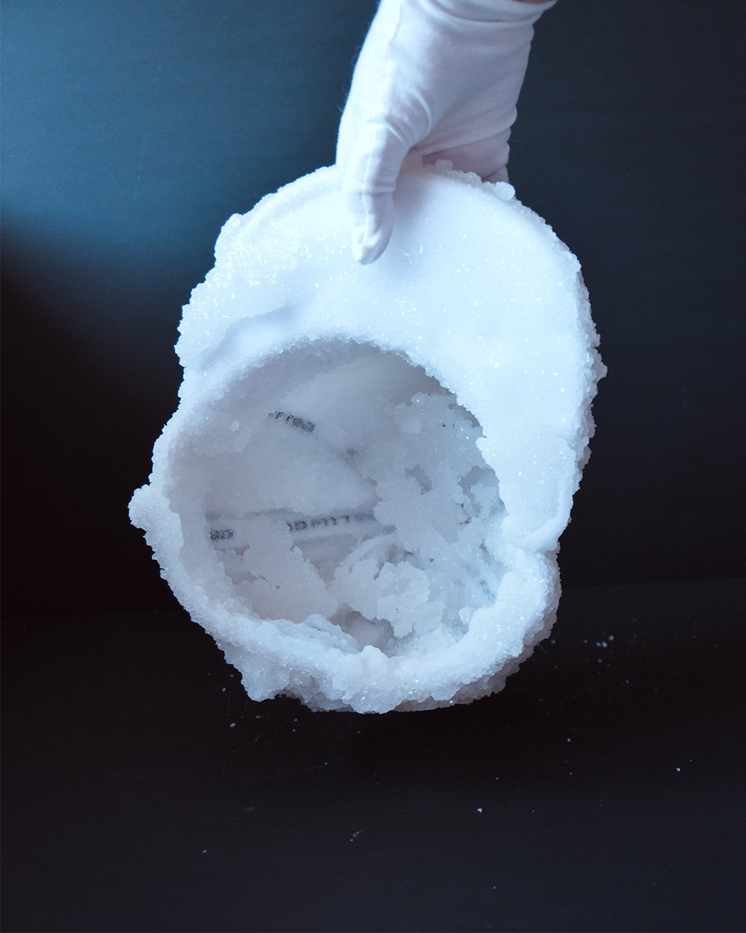 Person wearing white gloves holding a cap covered in crystals over a black surface with small crystal particles around.