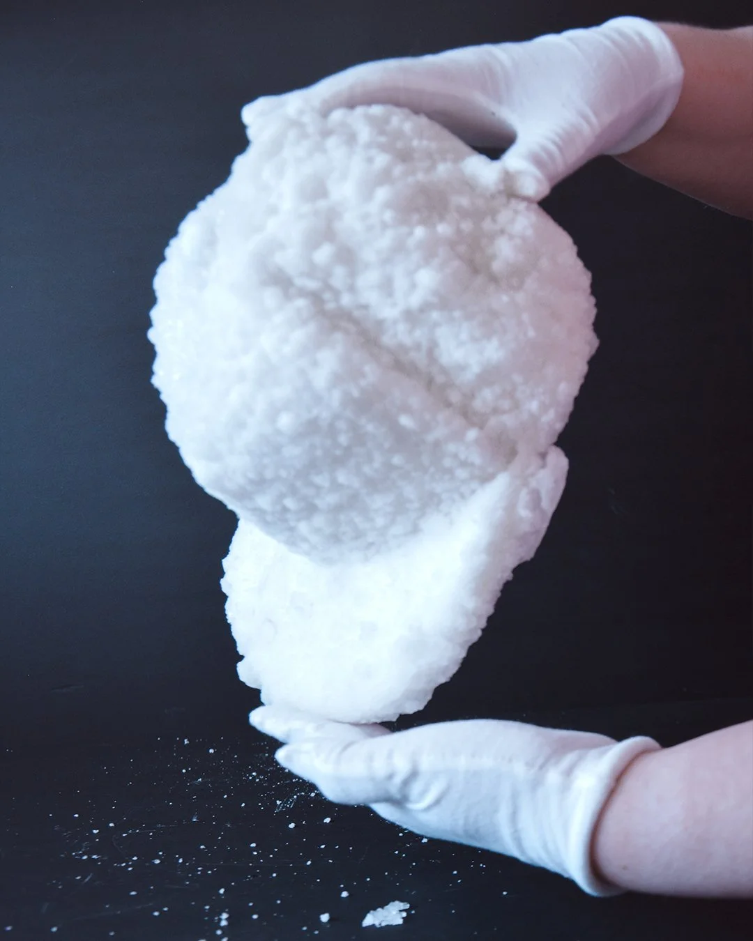 Person wearing white gloves holding a cap covered in crystals over a black surface with small crystal particles around.