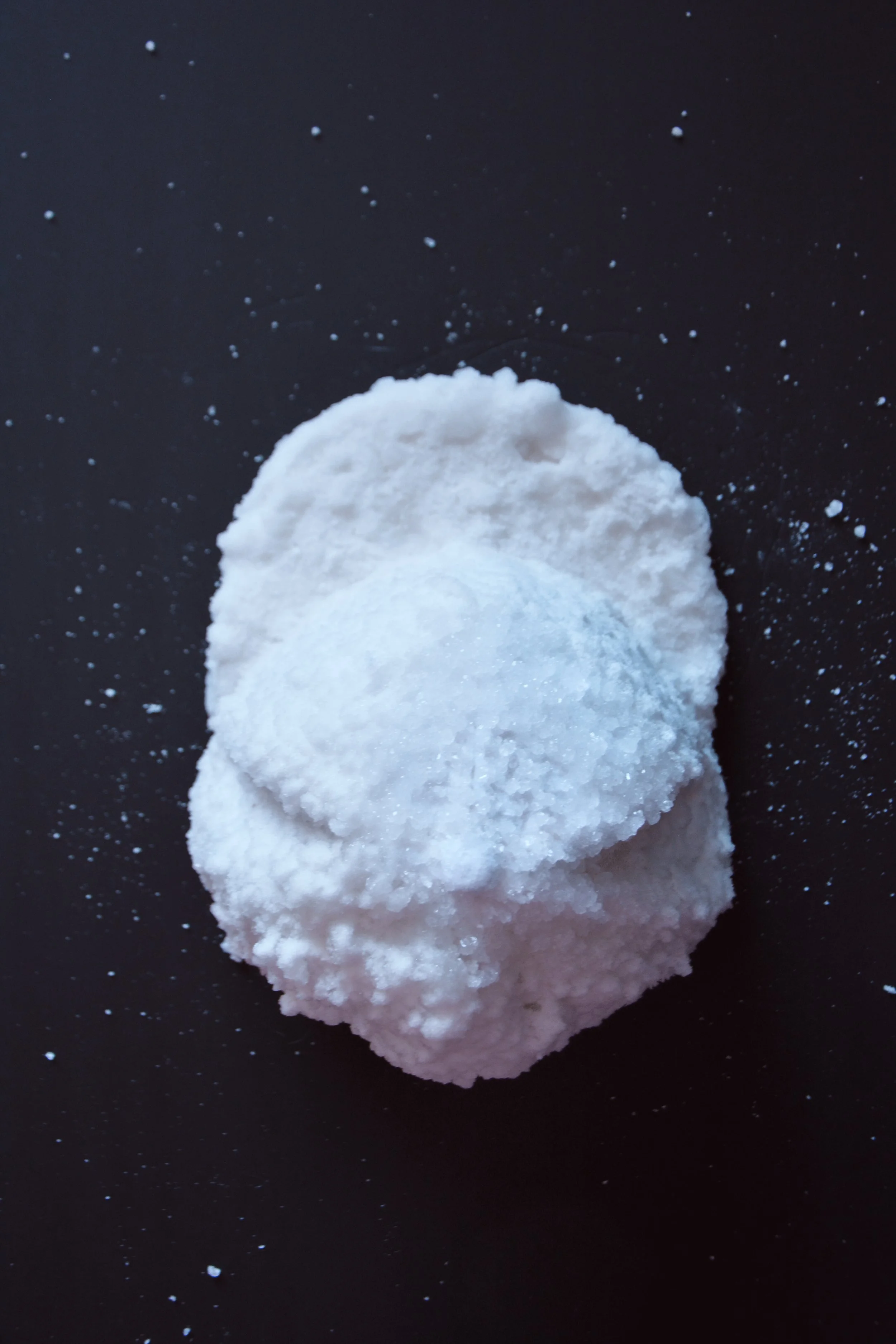 A white, rough-textured salt rock on a black background with salt particles scattered around it.