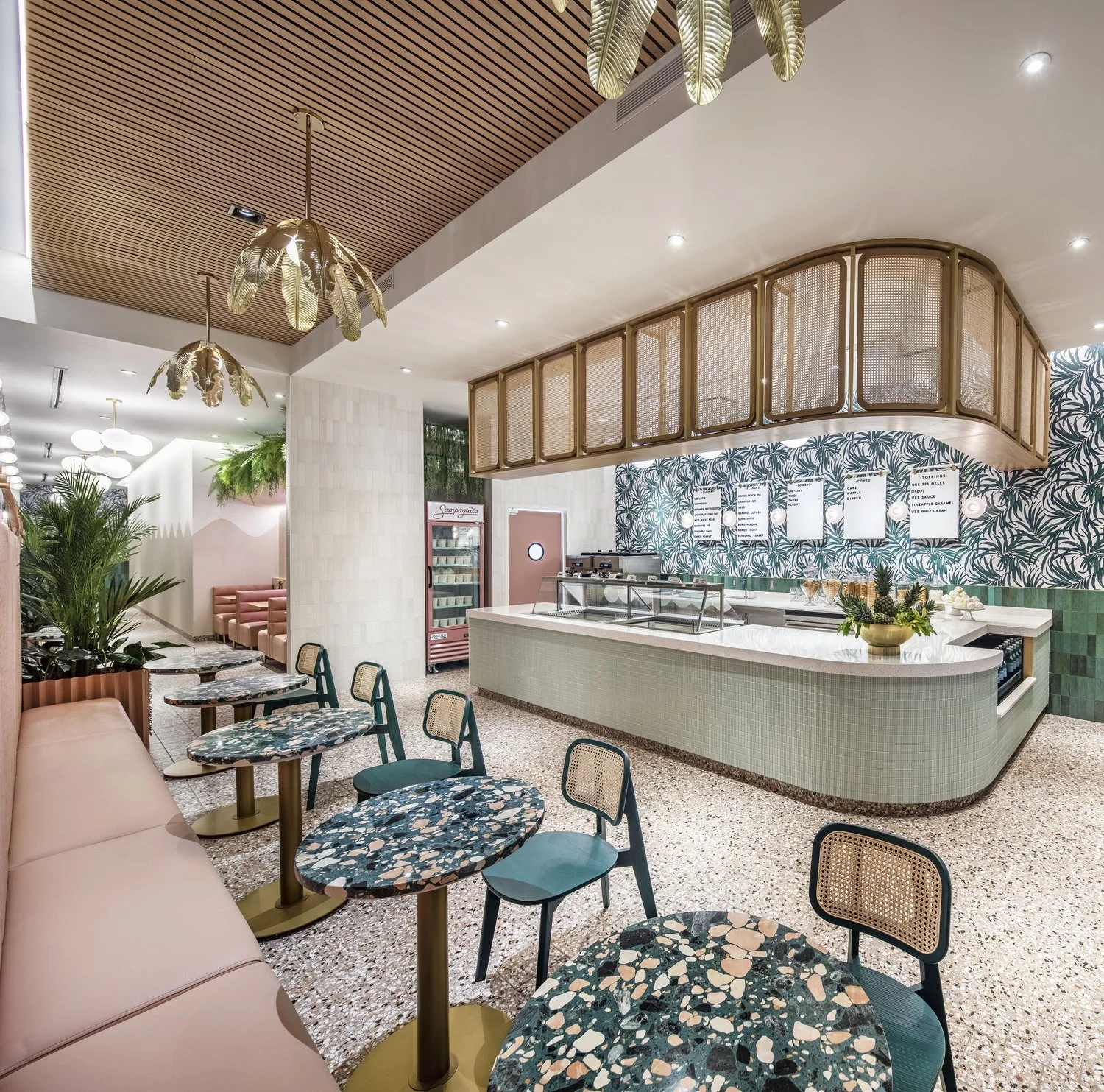 Interior of a modern cafe with a pastel green curved counter, decorative wallpaper with green palm leaves, and colorful terrazzo tables with matching chairs. Hanging gold pendant lights shaped like leaves and lush green plants decorate the space.