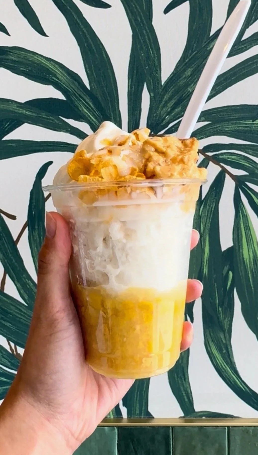 Hand holding a clear plastic cup of corn and vanilla soft cream ice cream and leche flan with a white straw against a leafy green plant background.