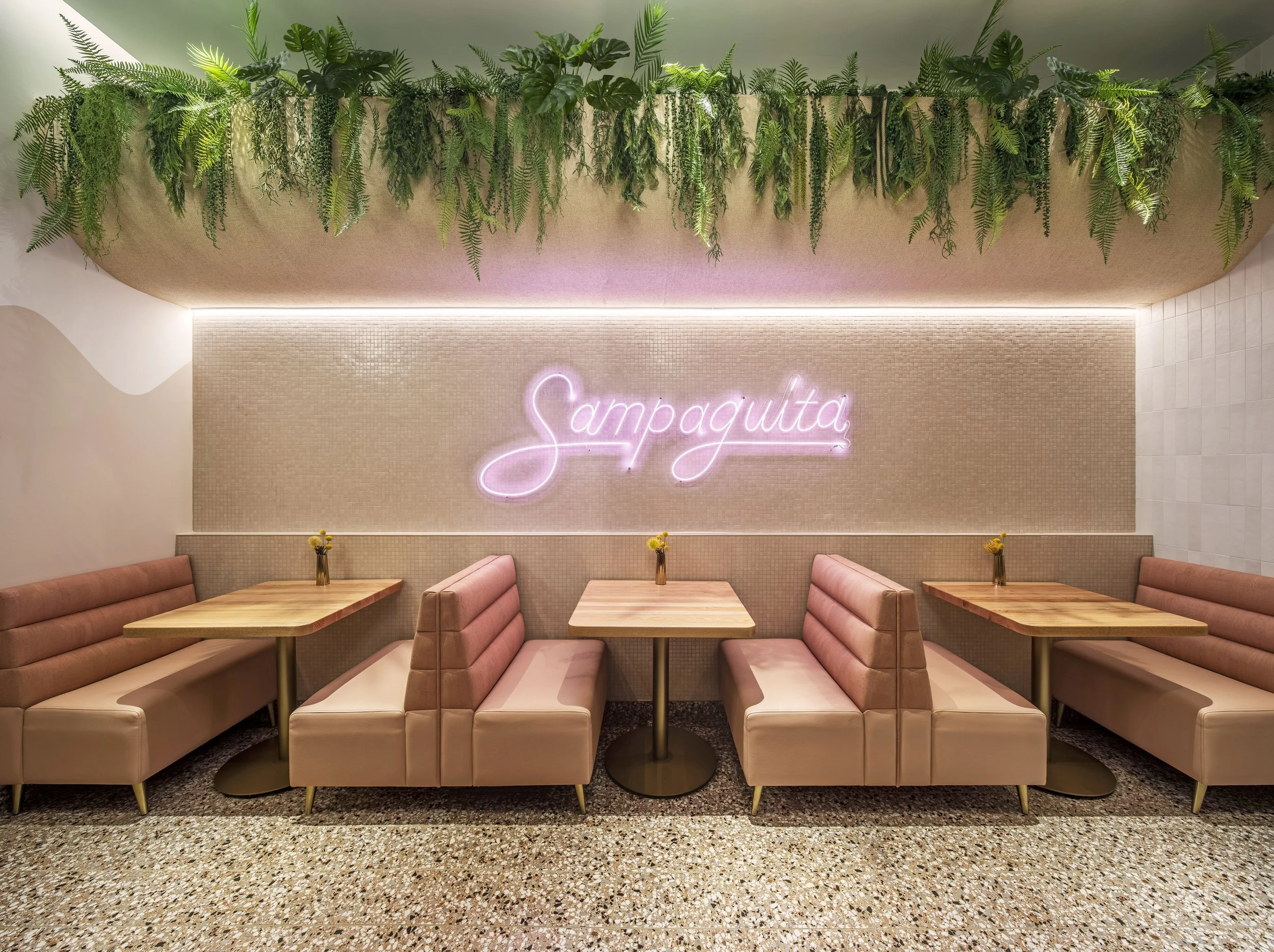 Interior of a restaurant or cafe with pink booths, wooden tables, small vases with yellow flowers, a textured beige wall with a pink neon sign reading 'Sampaguita,' and a green leafy plant decoration hanging from the ceiling.