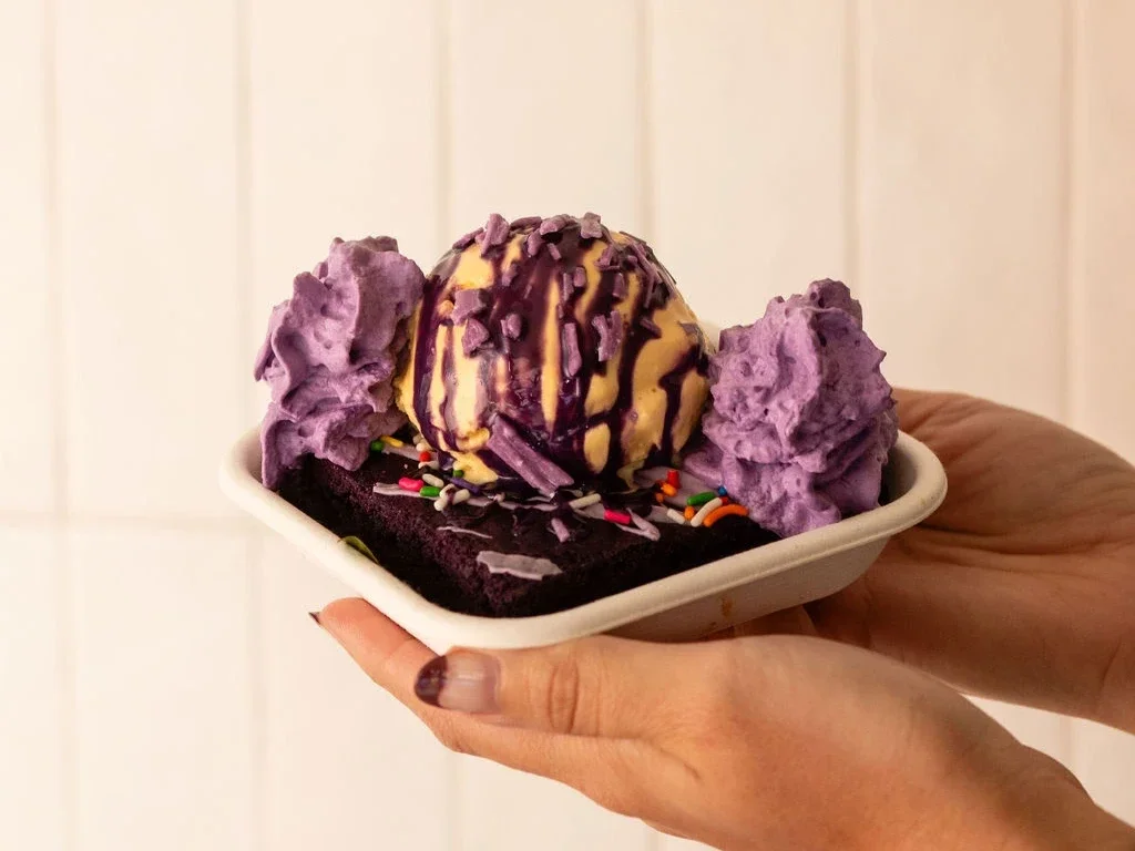 Warm homemade ube brownie topped with a scoop of ice cream, ube sauce, ube sprinkles, and ube whipped cream