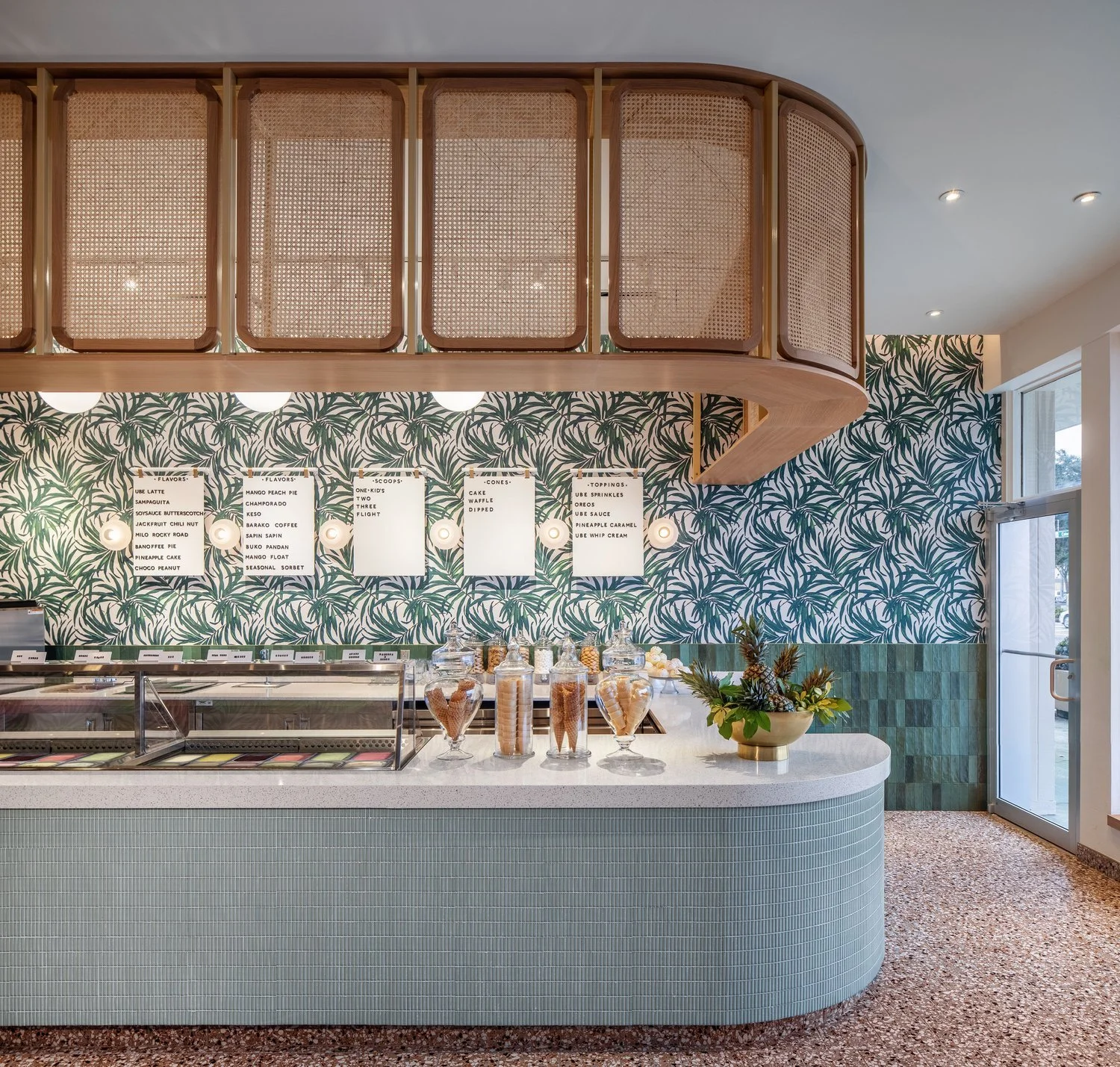Interior of a modern ice cream or frozen yogurt shop with a tropical themed wallpaper, glass jars of toppings and treats on the counter, and a curved light blue tiled front counter.