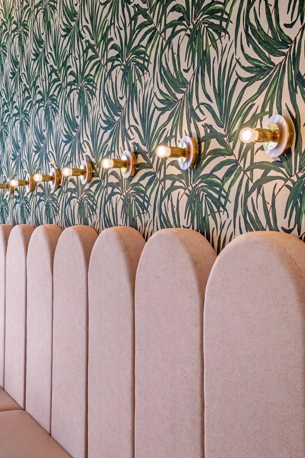 Pink upholstered headboard against a jungle-themed wallpaper with green leaves, illuminated by a row of wall-mounted light fixtures.