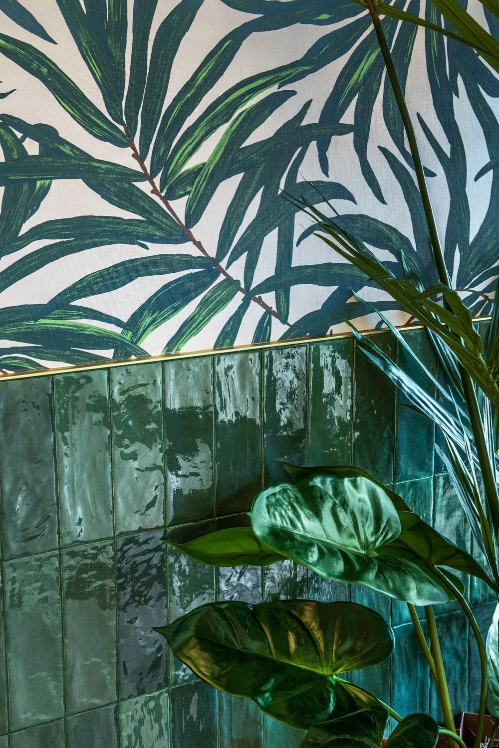 Green leafy plants in front of a wall with green tiles and botanical wallpaper with large green leaves.