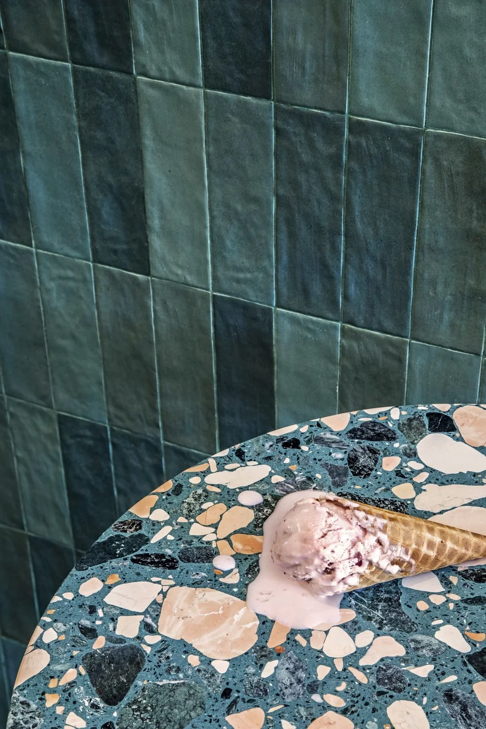 An ice cream cone resting on a multicolored terrazzo table with a green tiled wall in the background.