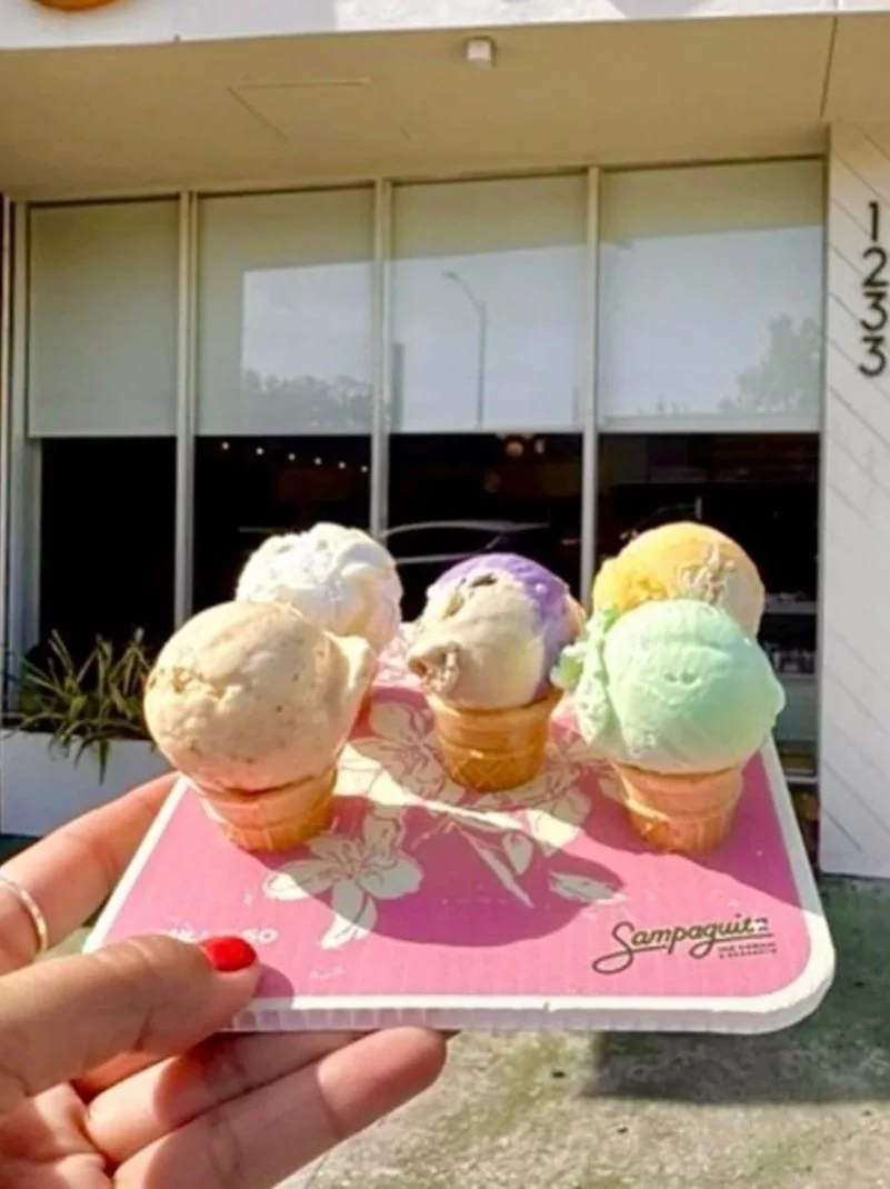 A hand holding a pink tray with five small ice cream cones, each with a scoop of different colored ice cream, in front of a building with large glass windows.